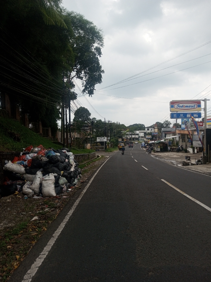 This image depicts a street scene in Kalchini, India, under an overcast sky. In the foreground, a substantial pile of refuse, primarily composed of black and white plastic sacks, rests on a dirt and grassy verge bordering a paved asphalt road. The road, featuring a solid white edge line on the left and a dashed white center line, extends into the distance, curving slightly to the right.
A single individual on a black scooter or motorcycle, wearing a helmet, is observed traveling away from the viewer in the left lane. Further down the road, a yellow three-wheeled vehicle is visible, along with other indistinct vehicles and people.
To the left of the road, a steep, vegetated embankment covered in dense green foliage rises, obscured by numerous utility wires strung between poles. The right side of the road is lined with various commercial structures. A prominent red and blue "Indomaret" sign is visible, along with smaller shops, stalls, and several individuals present on the roadside. Utility poles and overhead wires are also evident on this side. The background shows more buildings and trees, with hills under the overcast sky.