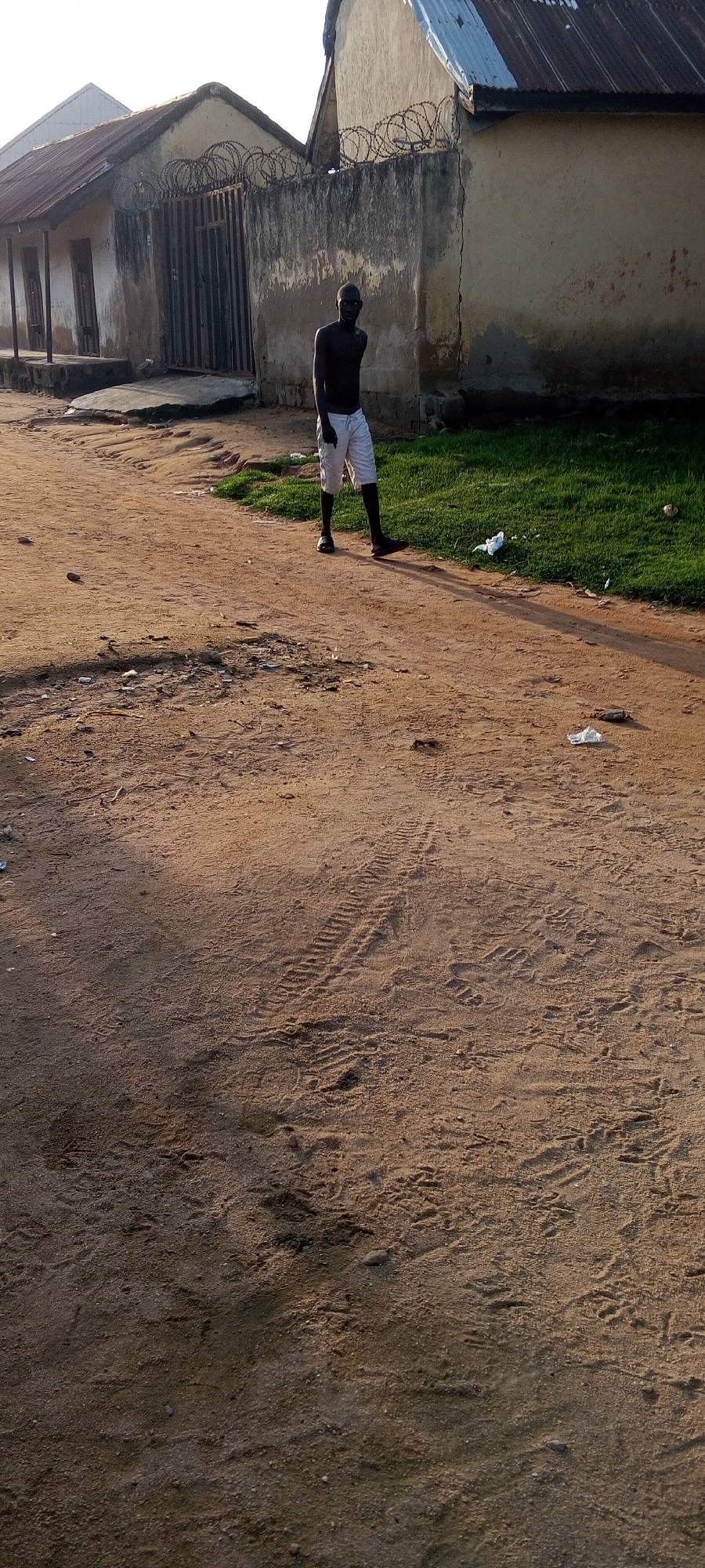 A young Black man stands on a dirt road in front of a weathered building with a metal gate topped with barbed wire. He is shirtless and wearing light-colored shorts and dark shoes. The ground is sandy with tire tracks visible. To the right of the man is a patch of green grass, and further on, a light-colored building with a corrugated metal roof. The sun is low in the sky, casting long shadows, suggesting it is either early morning or late afternoon. The scene appears to be in a rural or semi-urban area, possibly in Jalingo, Nigeria, given the context. The man's posture is relaxed, and he is looking towards the camera. There are some pieces of litter scattered on the ground and grass.