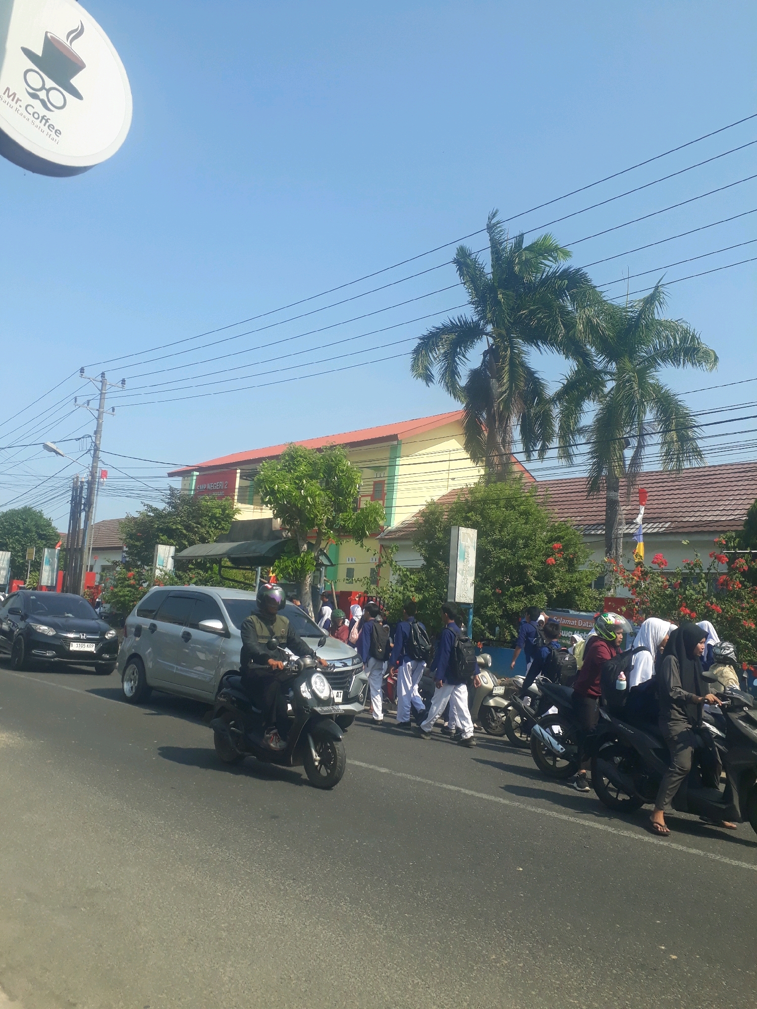 The image shows a street scene in Pangkalpinang, Indonesia, under a clear blue sky. A group of students, many wearing dark blue uniforms and white pants or skirts, are gathered on the sidewalk and crossing the street. Some are on motorcycles, while others are walking. A silver minivan and a black car are visible on the road, suggesting traffic. A building with yellow and green walls, identified as "SMP NEGERI 2 PANGKALPINANG" (State Junior High School 2 Pangkalpinang), stands in the background, flanked by palm trees and lush greenery with red flowers. A sign for "Mr. Coffee" is also visible in the upper left corner. The scene appears to be during daytime, possibly around school dismissal time, as indicated by the presence of students.