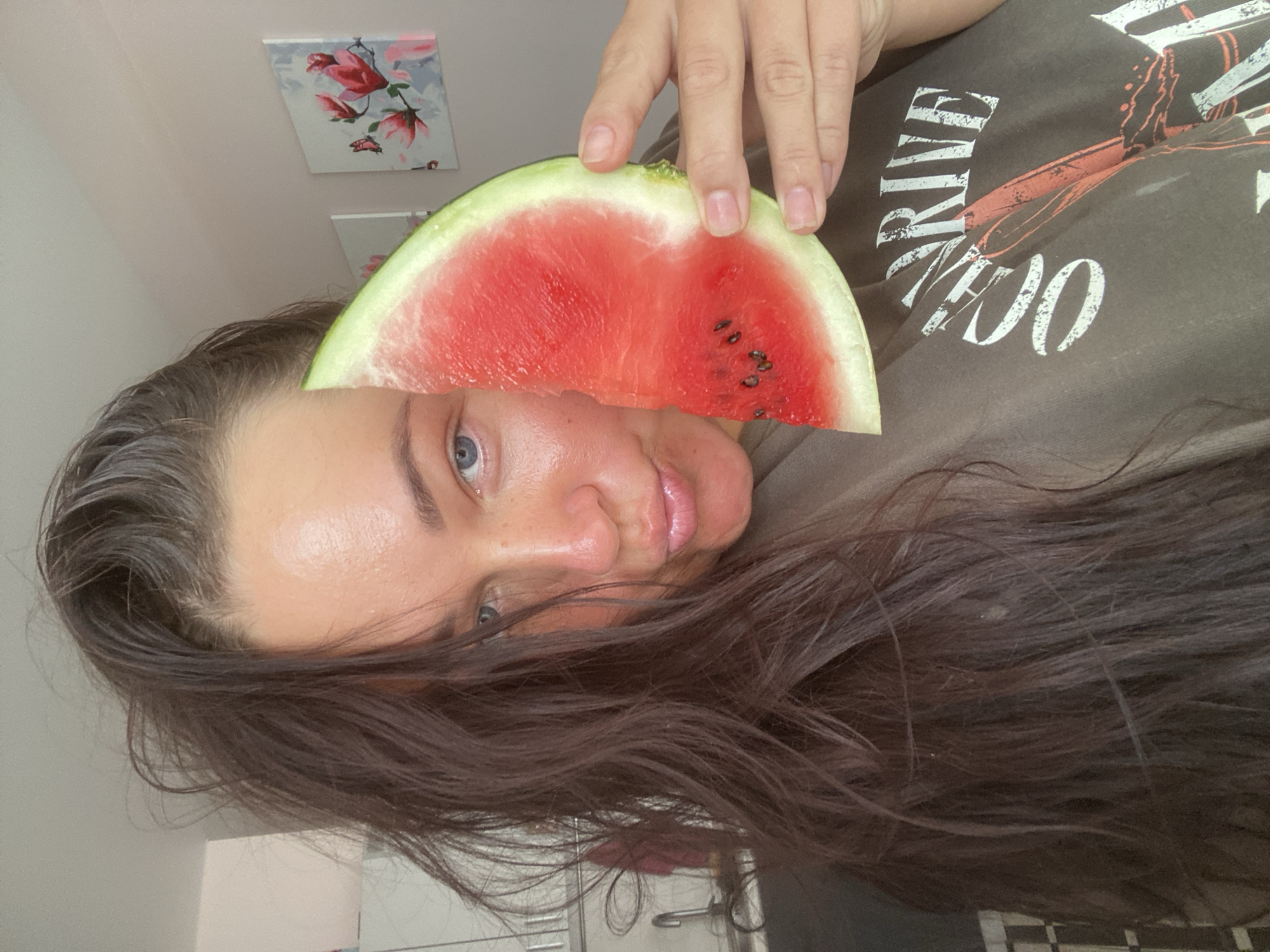 A young woman with long, dark hair is holding a slice of watermelon in front of her face. She is wearing a dark t-shirt with a graphic print. The background appears to be an indoor setting, possibly a home, with a kitchen sink visible on the left and framed artwork on the wall to the right. The image does not provide any information about the city of Sochi, Russia.