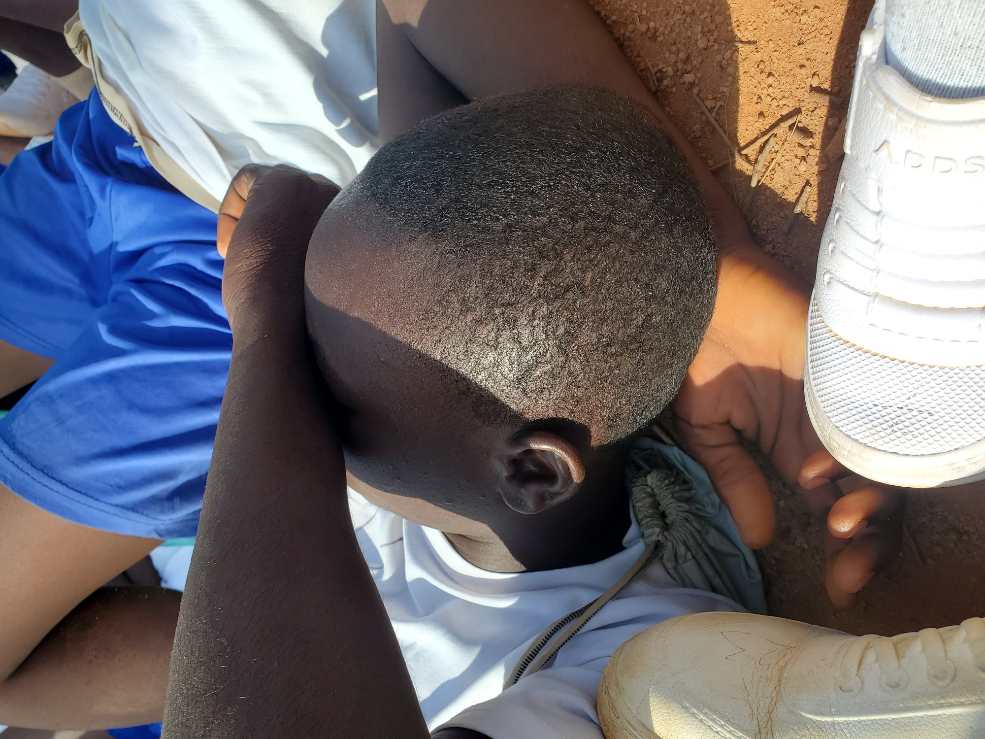 The image shows a close-up, overhead view of a person lying down, with their head tilted to the side. They are wearing a white t-shirt and blue shorts. Their arms are bent, and their hands are resting on their head, possibly to shield their eyes from the sun. The person's skin tone is dark, and their hair is short and dark. White sneakers are visible in the foreground and to the right of the frame. The ground beneath appears to be sandy or dusty, with some small sticks scattered on it. The lighting suggests it is daytime, with strong shadows cast by the person's head. The scene seems to be outdoors, possibly in a rural or open area given the ground texture. There is no discernible text in the image.