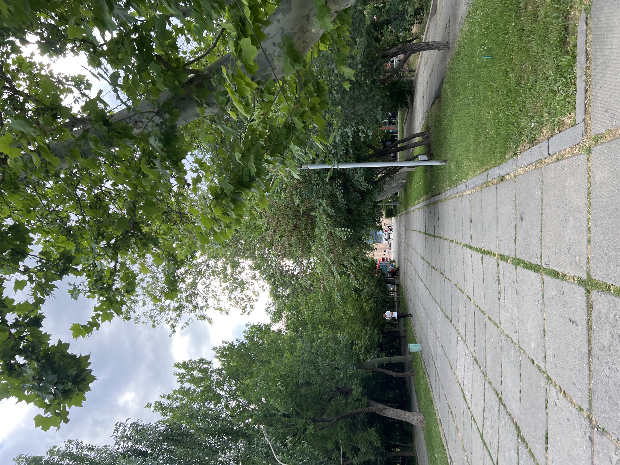 The image depicts a pedestrian walkway in a park-like setting in Yerevan, Armenia. The walkway is paved with large, square concrete slabs, with grass growing in the gaps between them. Trees line both sides of the walkway, with lush green foliage. In the distance, several people can be seen walking and sitting, suggesting a public space. The sky above is partly cloudy. The overall impression is one of a pleasant, tree-lined urban pathway.