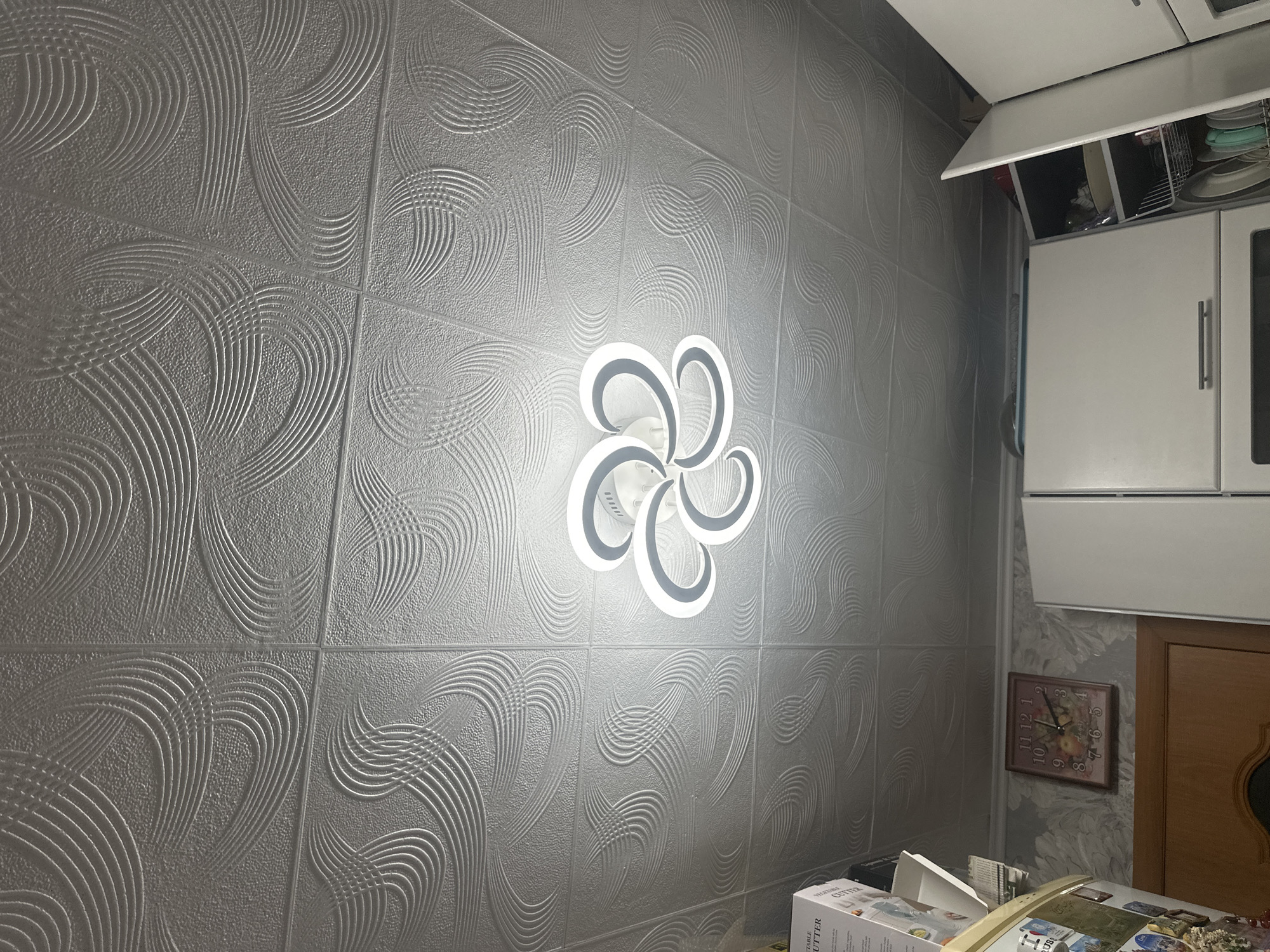 An interior view shows a white embossed ceiling with a swirling pattern. A modern white light fixture with curved arms and LED lighting is centrally mounted on the ceiling. Below the ceiling, on the wall, a framed analog clock with a floral design is visible. To the left, a refrigerator with various magnets and items on its door is partially visible, as well as a box labeled "VEGETABLE CUTTER." To the right, white kitchen cabinets with open doors reveal shelves with dishes and other kitchenware. The image depicts a room, likely a kitchen, within a dwelling in Загородный, Russia.