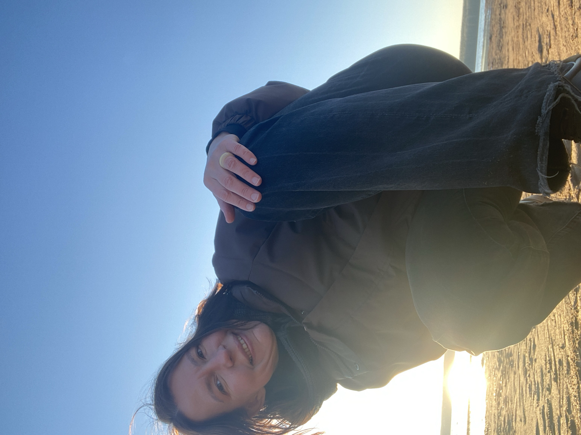 A woman is crouching on a sandy or concrete surface. She is wearing a brown jacket and black jeans with frayed hems. Her hands are clasped around her knees. The background consists of a clear blue sky and a bright light source, likely the sun, creating a strong glare. The location is not explicitly stated to be the city of Chaikovsky, Russia, in the image itself. Therefore, information on the location cannot be definitively extracted from the image alone.