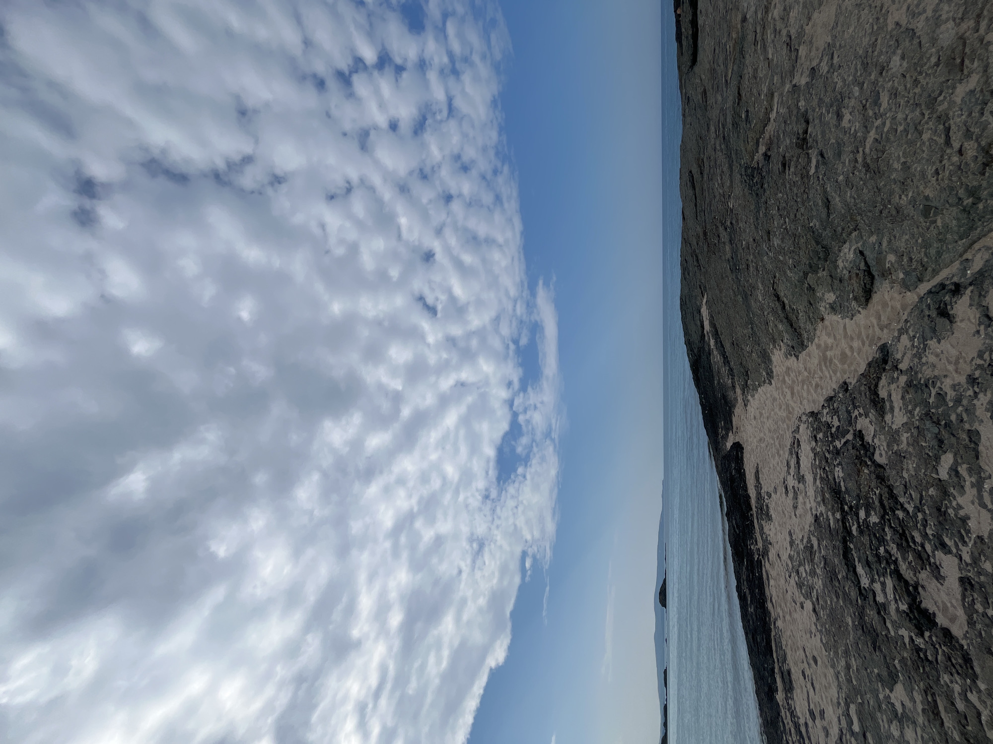 The image depicts a rocky shoreline at the sea. The foreground is dominated by dark, rough rocks interspersed with sandy patches. A sandy path winds its way through the rocks, leading towards the water. The sea is calm and a light blue, stretching out to the horizon where distant landmasses or islands are visible. Above the sea, the sky is filled with a dramatic array of white and gray clouds, creating a textured pattern against the clear blue sky. The overall scene evokes a natural, coastal environment. The location is the city of Lozenets, Bulgaria.