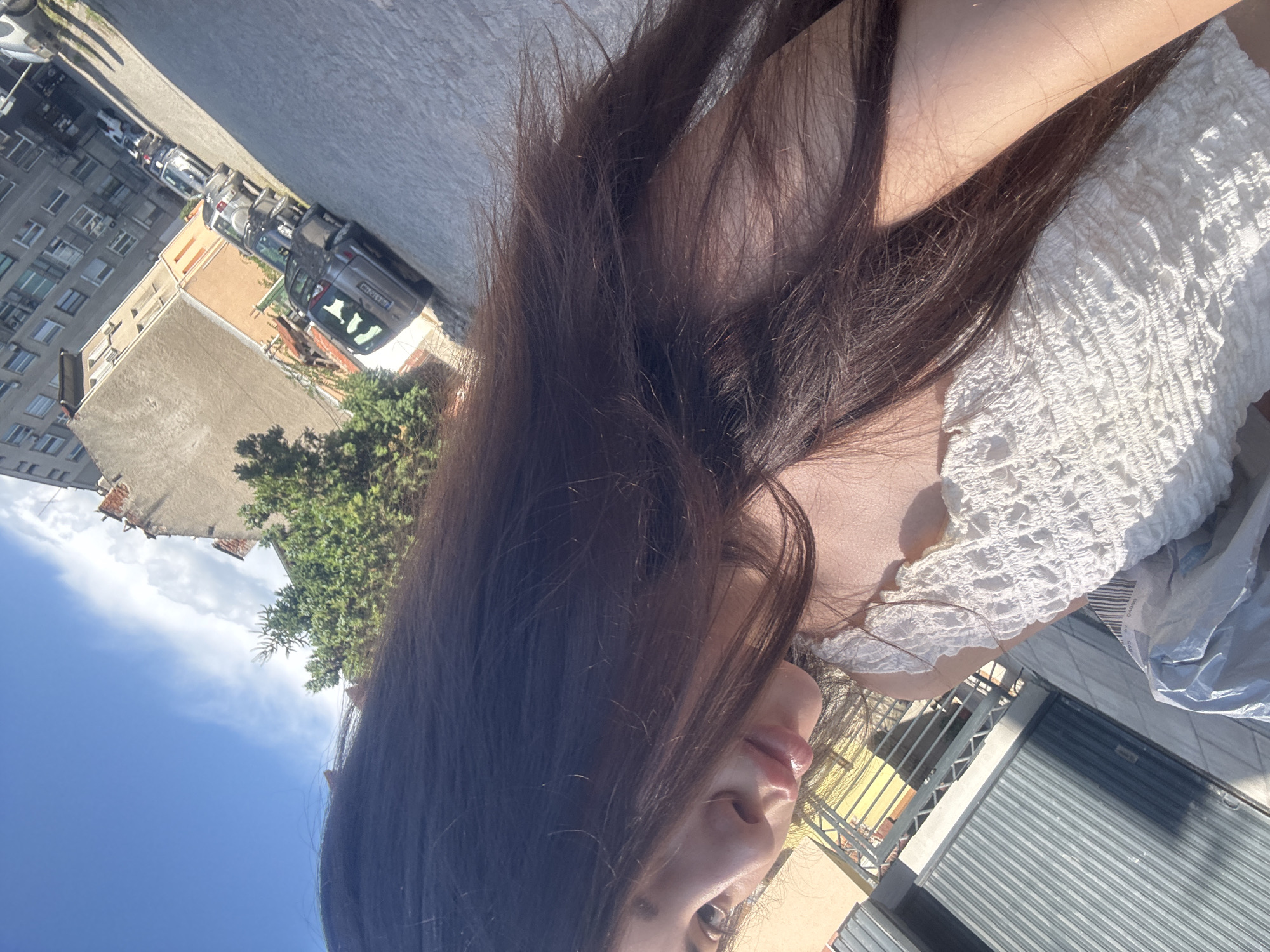 The image, when correctly oriented, presents a close-up, low-angle perspective of a person outdoors on a bright, sunny day in an urban setting. The foreground is dominated by the person's long, dark brown hair, which partially obscures their face, revealing only their lips, chin, and a section of their eye. They are wearing a textured, white smocked or ruffled top.
In the background, an urban street scene unfolds under a clear blue sky with scattered white clouds, indicating sunny weather, likely morning or afternoon. Parked cars line a paved street, and behind them stand multi-story buildings; one has a light brown/orange facade, and another is grey with visible windows. Green foliage from trees and bushes is interspersed within the urban landscape.
Notable details include partially visible Bulgarian license plates on the cars: one reads "BX 4104" (indicating Vratsa region) and another "CB 64" (indicating Sofia region). A grey corrugated metal rolling shutter, possibly a store entrance, is visible in the lower right, alongside a crumpled white plastic bag. The scene captures a casual moment in an urban environment identified as Plovdiv, Bulgaria.