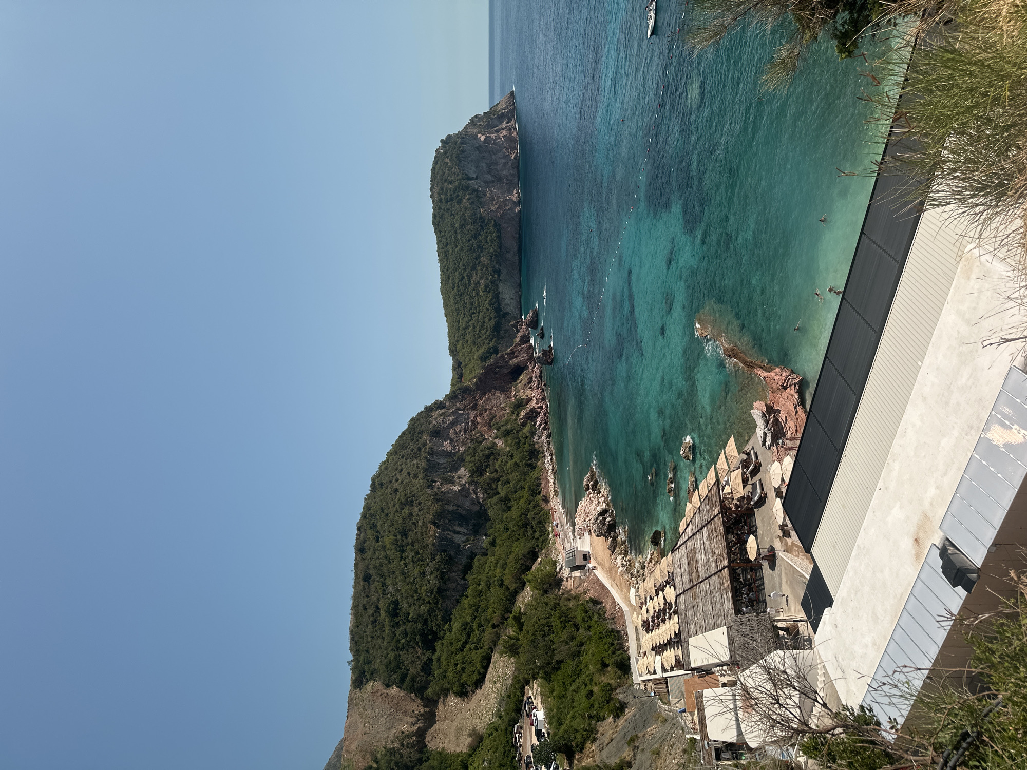 The image captures a coastal scene in Sveti Stefan, Montenegro. The foreground features a multi-level structure with flat roofs, some covered in dark material and others appearing as concrete. Beyond this structure, a beach area is visible with numerous rows of closed umbrellas and lounge chairs. Adjacent to the beach, the turquoise waters of the Adriatic Sea are clear, revealing submerged rocks. A small, rocky cove is present to the left, and further out, a larger, tree-covered headland juts into the sea, with another prominent cliff face further in the distance on the right. Several people are visible swimming in the water near the beach. A small white boat is anchored in the sea on the far right. The background is dominated by a steep, tree-covered mountainside. The sky is a clear, bright blue. A parking area with several cars is located on a slope above the beach on the left side of the image.