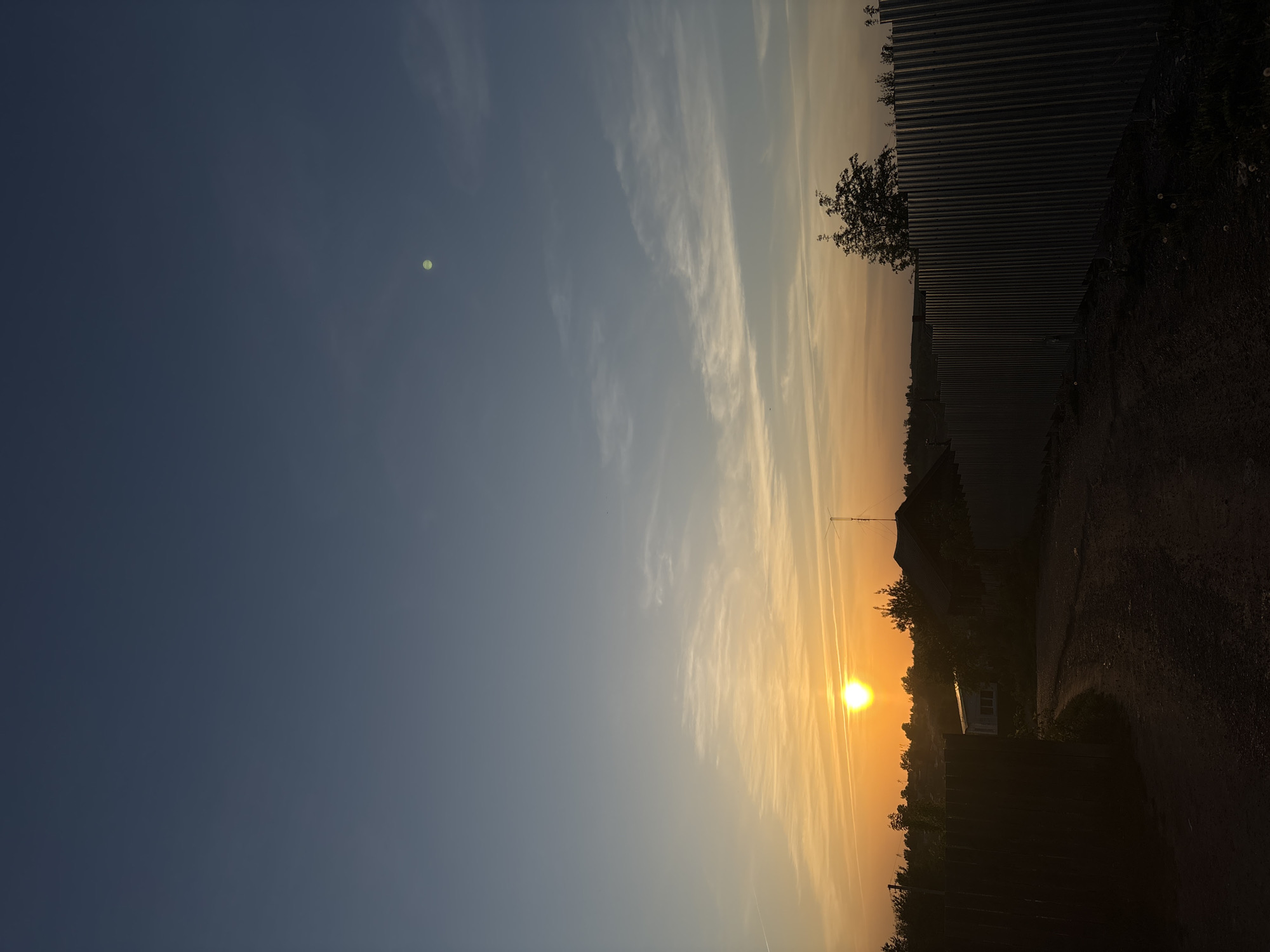 The image captures a sunset scene in Фоки, Russia. The sun is positioned near the horizon, emitting a bright, orange glow. Wispy clouds are present in the sky, transitioning from blue to orange hues. In the foreground, a metal fence and trees are silhouetted against the sunset. A building with a visible window is also part of the shadowed landscape. A road is also visible along the right side of the image.