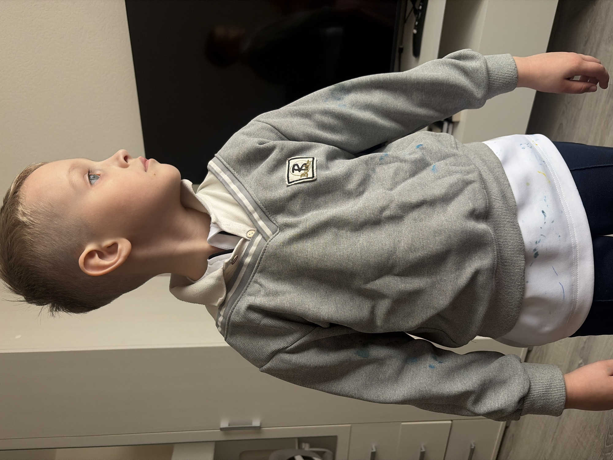 A young boy is pictured indoors. He is wearing a grey jacket over a white collared shirt and dark blue pants. The jacket has an emblem on the left chest. His hands are visible. The background includes a white cabinet, a dark television, and light flooring. There is no indication of the location being Saint Petersburg, Russia.