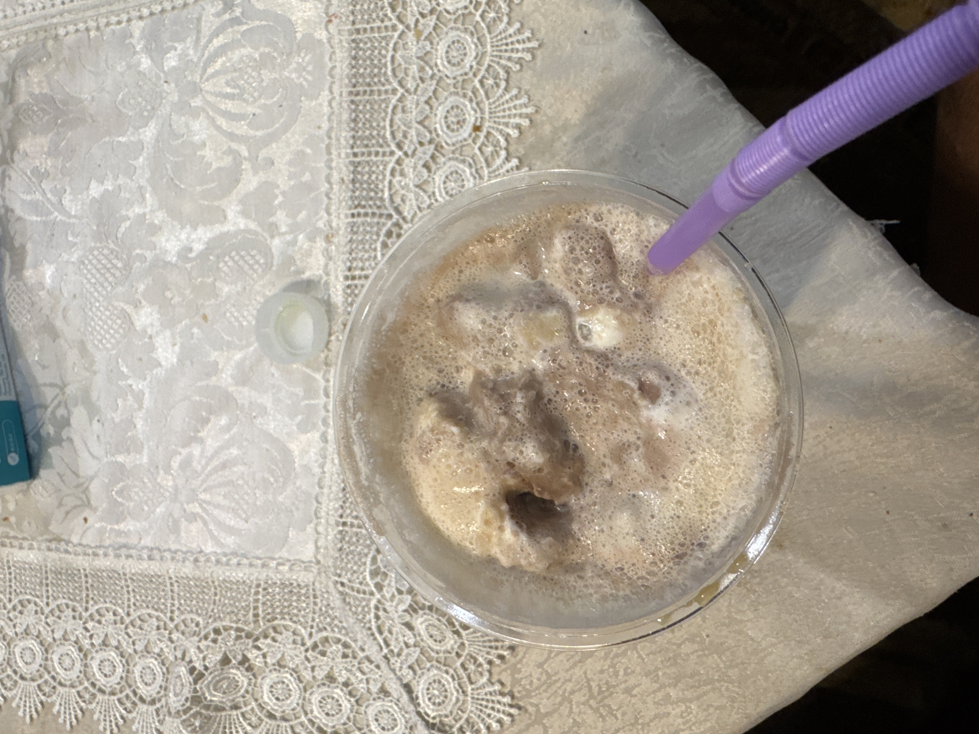 An overhead shot shows a plastic cup filled with a frothy, light brown liquid, likely a milkshake or smoothie, with chunks of a darker substance, possibly ice cream or frozen fruit, visible within. A purple flexible straw is inserted into the drink. The cup is placed on a white surface, possibly a tablecloth or placemat, which features an intricate floral lace pattern along its border. Next to the cup, on the left, is a small white cap, seemingly detached. The background is a mix of white lace and a plain, off-white surface. This scene is indoors, potentially in Yerevan, Armenia.