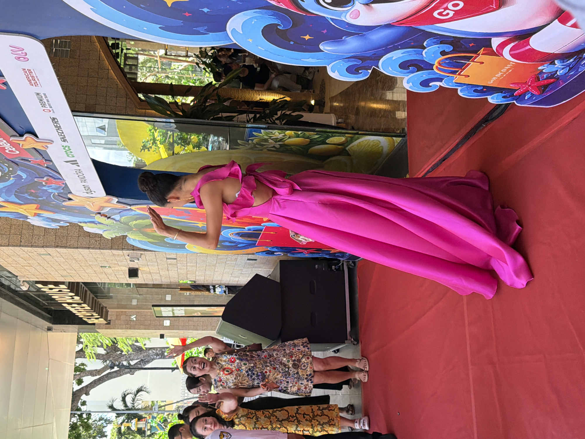 In Nha Trang, Vietnam, on a red carpet, a woman in a vibrant pink gown with a cutout back and ruffled sleeves poses with her hand raised. Several women in casual attire stand behind her, some waving. In the background, a building with a sign reading "THE COFFEE HOUSE" is visible. A large, colorful backdrop featuring waves, stars, and fruit is also present, along with a display of large black speakers. The scene appears to be an outdoor event, possibly a promotion or celebrity appearance, given the backdrop and crowd.