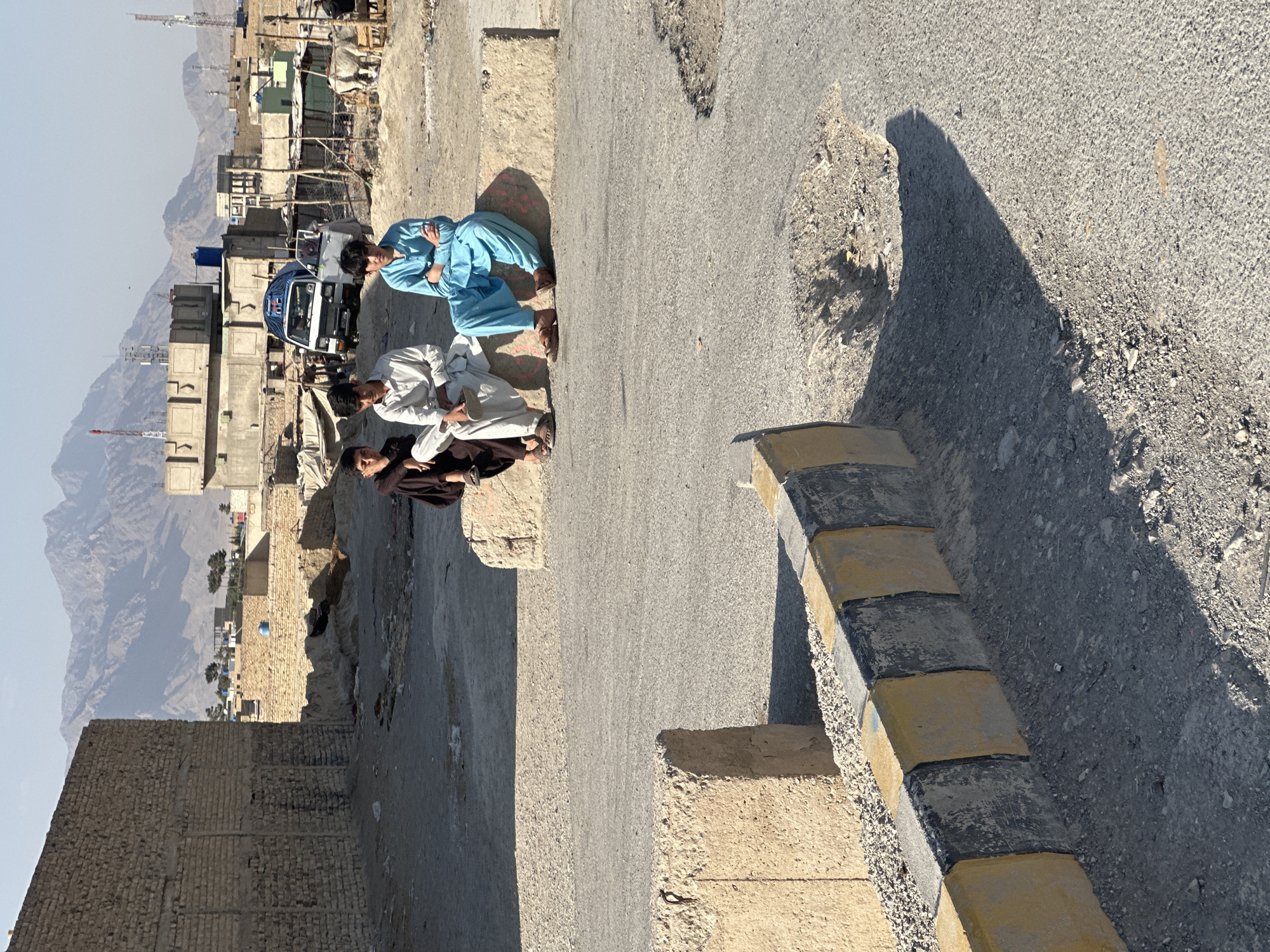 Three young males are seated on a concrete barrier in a dusty, urban environment. Two of the males are wearing traditional shalwar kameez, one in white and the other in a light brown garment. The third male is wearing a bright blue shalwar kameez. Behind them, a white van with a blue canopy is parked, alongside other buildings constructed from brick and corrugated metal. In the distance, mountains are visible under a clear sky. The foreground features a paved road with a curb marked with alternating yellow and black stripes, and a large pothole. This scene is characteristic of a developing area within Quetta City Tehsil, Pakistan, likely a street or public space affected by ongoing construction or disrepair, under the typical climate conditions of the region.