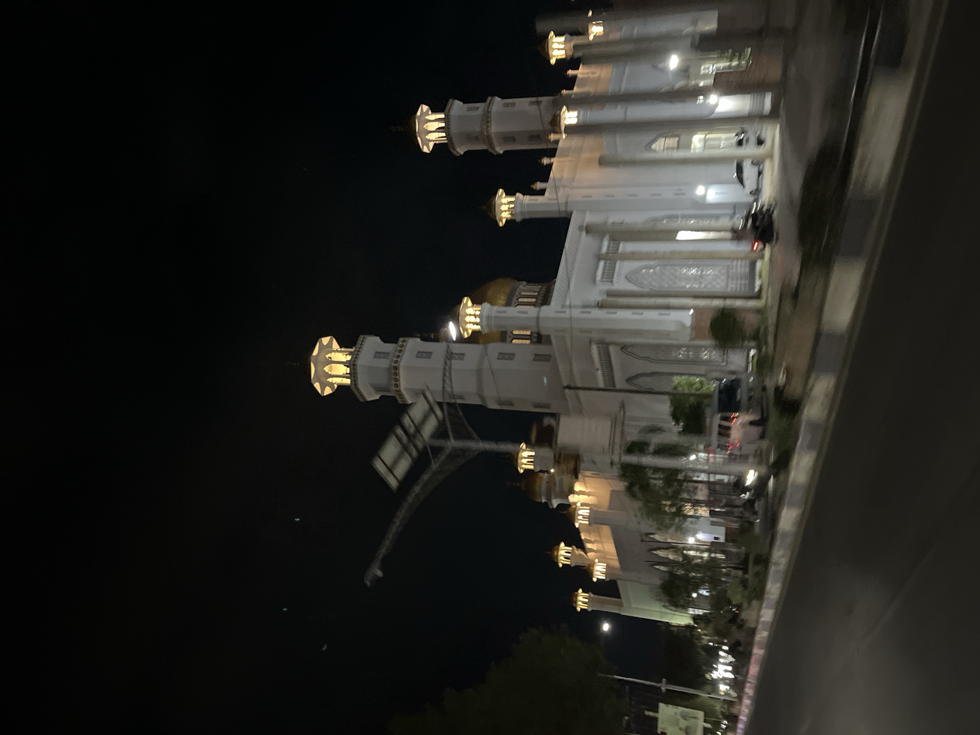 This image depicts the Masjid Agung Langsa in Langsa, Indonesia, at night. The mosque is illuminated, showcasing its white facade and multiple minarets topped with golden domes. The architectural style is traditional Islamic, with arched windows and decorative detailing. In the foreground, a street is visible, with a road and a sidewalk. Several vehicles, including cars and a van, are parked or driving on the street. Trees and greenery line the sidewalk in front of the mosque. The overall scene suggests a public space in an urban environment, with the mosque serving as a prominent landmark. The sky is dark, indicating it is nighttime, with a few faint lights visible in the distance, possibly streetlights or lights from other buildings.