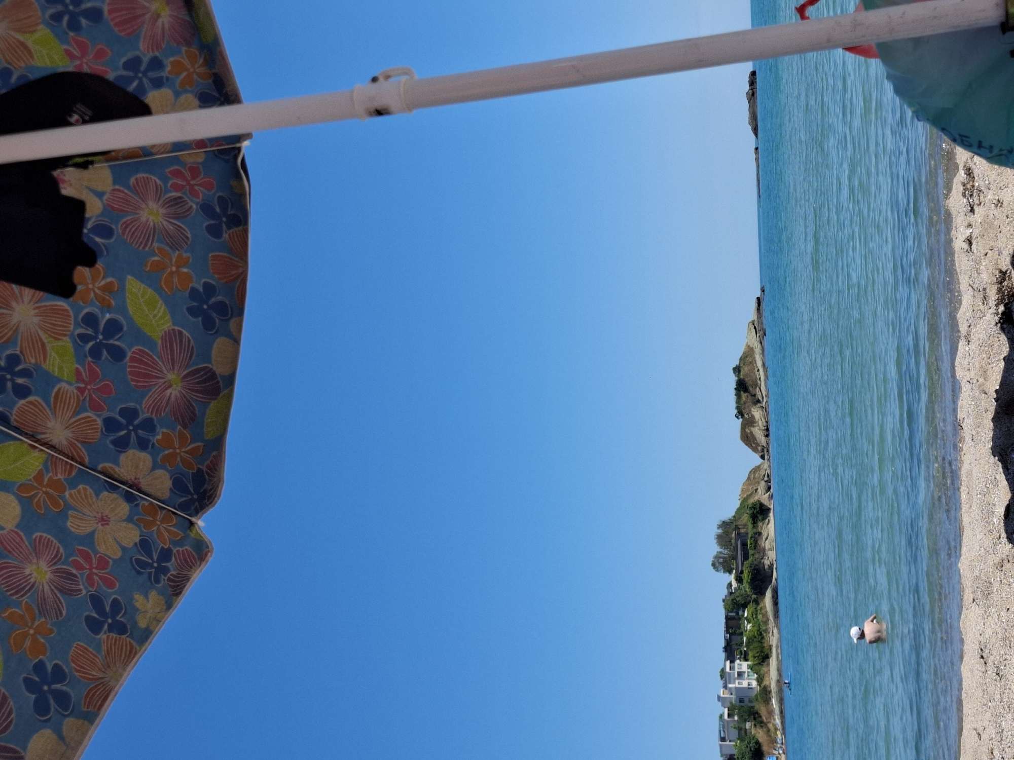 The image depicts a sunny day at a beach. The foreground shows a sandy shore with some seaweed. In the water, a child wearing a white cap is standing and facing away from the camera. Further out in the sea, there are some small islands and a distant coastline with buildings. An umbrella with a floral pattern provides shade in the upper left corner of the image, and a white umbrella pole is visible on the right side. The sky is clear and blue, indicating good weather.