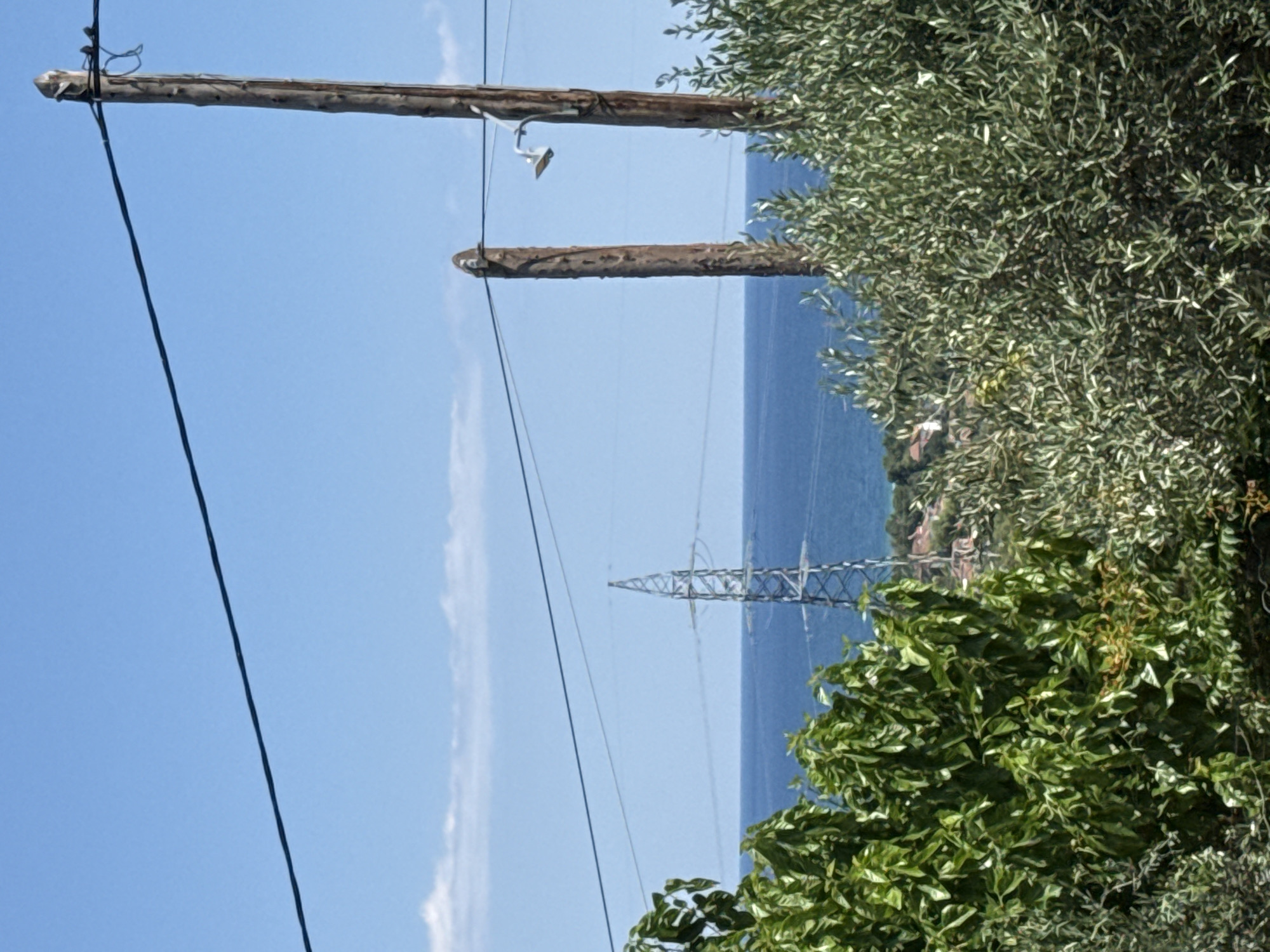 In Asprovalta, Greece, on a clear, sunny day, the image captures a scenic view overlooking the sea. In the foreground, lush green trees partially obscure the view of the landscape. Two wooden utility poles stand prominently, with power lines stretching across the frame. One pole is adorned with a light fixture. In the middle ground, a lattice-like power pylon stands tall against the horizon. Beyond the pylon, the deep blue sea stretches out, meeting the light blue sky with a few wispy white clouds. The distant shoreline is dotted with buildings and more greenery, suggesting a coastal town or village. The overall scene depicts a tranquil, natural environment integrated with man-made infrastructure.