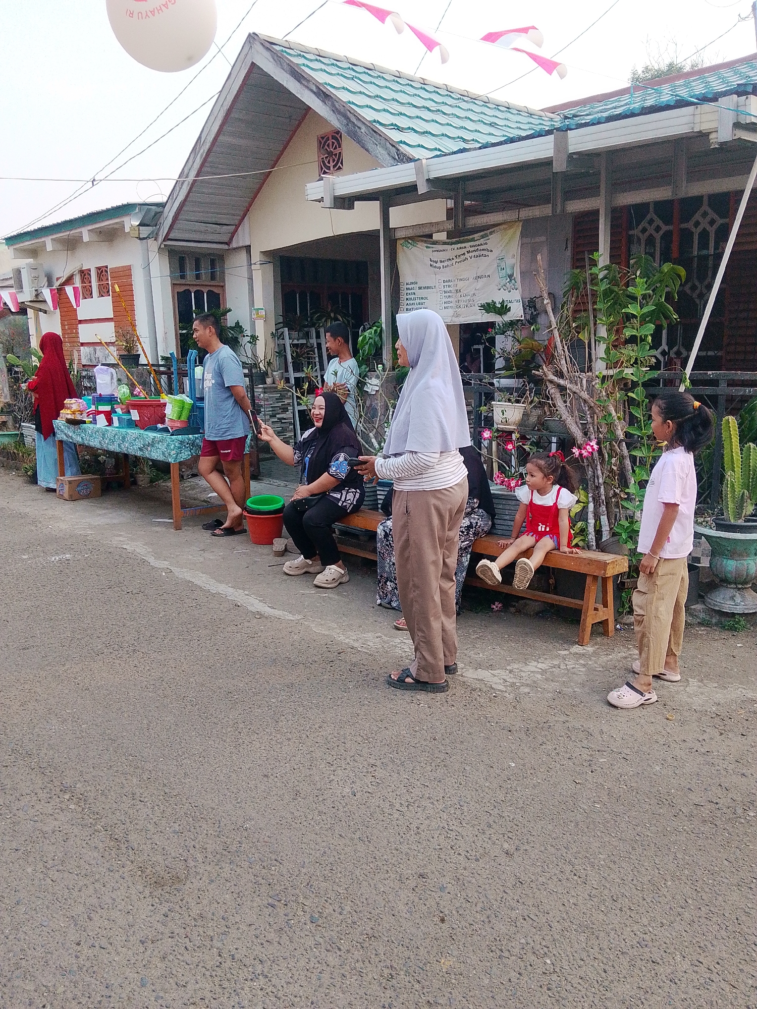 The image depicts a street scene in Barabai, Indonesia, with several people gathered. In the foreground, a paved road is visible. To the left, a table is set up with various items, possibly for sale or display, including containers and a box. A woman in a red headscarf stands near the table. A man in a blue t-shirt and red shorts stands near the table, facing away from the camera. Behind them, a house with a green roof and white exterior is visible, decorated with Indonesian flags.
Further to the right, a group of people are seated and standing. A woman in a black headscarf and patterned top is sitting on a wooden bench, holding a mobile phone and appearing to take a photo. A younger girl in a red dress is also sitting on the bench. Another girl, wearing a pink t-shirt and brown pants, stands nearby. An adult woman wearing a grey headscarf and brown pants stands to the left of the bench, looking towards the right. A man and a child are also present, partially obscured. Plants and decorative items are visible in front of the house. In the background, a large white balloon with text is visible in the sky. The overall scene suggests a local gathering or event.