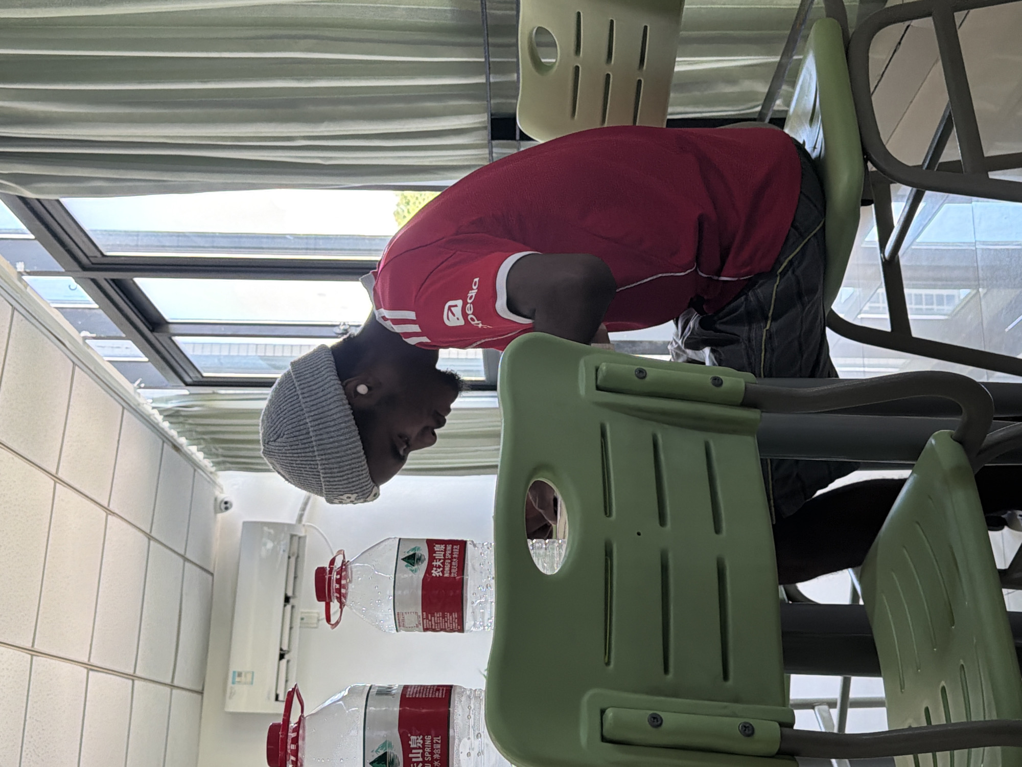 The image shows a young Black man wearing a grey beanie and a red t-shirt with the Expedia logo, seated at a table in what appears to be a classroom or common area. He is looking down, seemingly engaged in an activity on the table. Two large plastic bottles of Nongfu Spring water are visible on the table. The environment features light-colored tiled ceiling, an air conditioning unit mounted on the wall, and large windows with light green curtains. In the foreground, two green plastic chairs are prominent. The scene is likely within a facility in Jiangjin, China, given the presence of Nongfu Spring water bottles, a popular Chinese brand.
