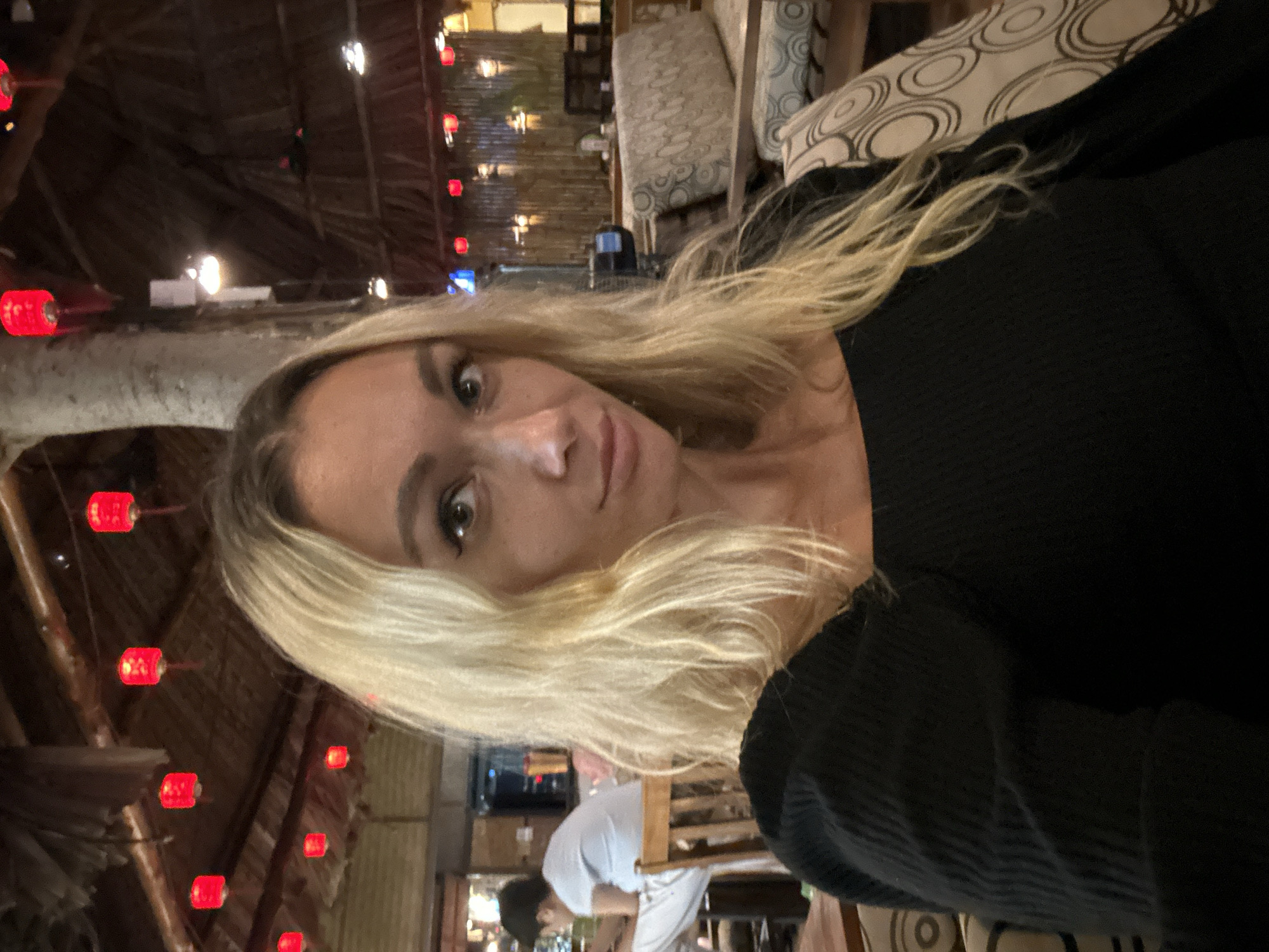 The image is a selfie taken by a woman with blonde hair and light skin, wearing a black top. She is seated indoors in what appears to be a restaurant or lounge area. The background shows a thatched roof, bamboo walls, and red lanterns hanging from the ceiling, characteristic of a Southeast Asian setting. There are also upholstered chairs and a table visible. To the left, a person in a light blue shirt and white pants is seated at a wooden chair, looking down at something in their lap. The woman in the foreground is looking directly at the camera. The overall ambiance suggests a casual, dimly lit environment. The location is specified as Phu Thuy Ward, Vietnam.