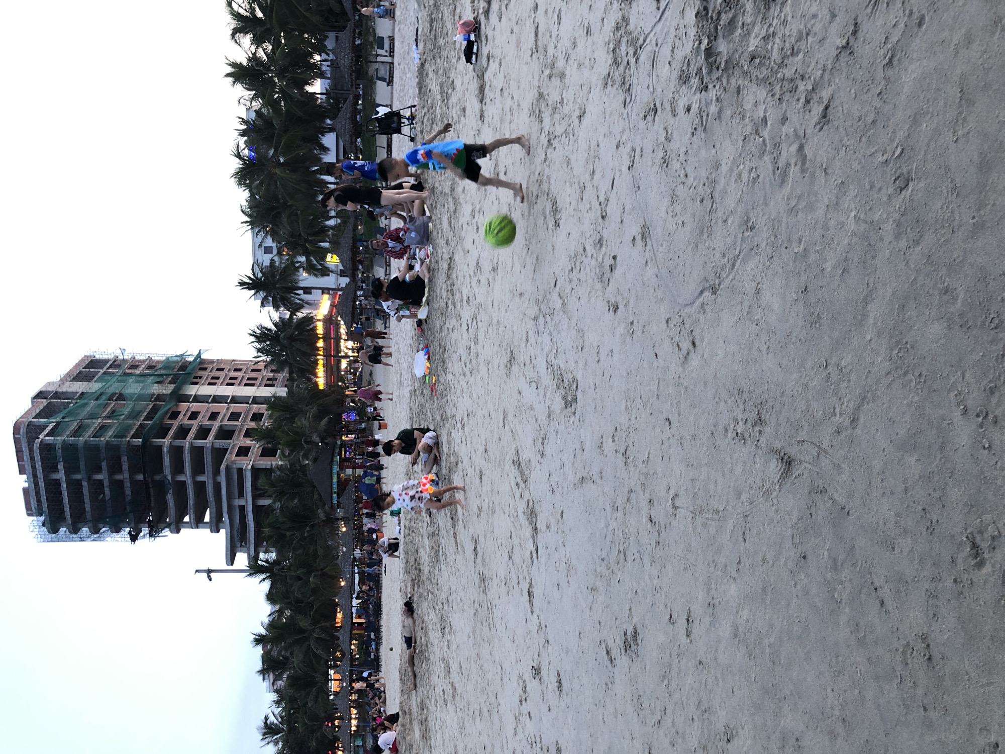 The image captures a beach scene in Da Nang, Vietnam, with a partially constructed building dominating the background. The building, covered in green netting, appears to be a high-rise hotel or apartment complex under development. In the mid-ground, several people are visible on the sandy beach.
In the foreground, a young boy in a blue and green t-shirt and black shorts is kicking a large, green ball. His body is angled, indicating he is in the process of kicking. To the left and slightly behind him, a young girl in a white, patterned dress is walking and holding a colorful object.
Further back, a group of people are sitting on a blanket on the sand, some of them appearing to be eating or picnicking. A man in a dark t-shirt and shorts sits cross-legged, looking at his phone. Another man lies down on the sand next to him, wearing black shorts.
In the distance, palm trees line the beach, interspersed with low-rise buildings, some of which appear to be restaurants or shops with awnings. Umbrellas are set up on the beach, providing shade. The sky is overcast, suggesting it might be late afternoon or early evening. The sand appears somewhat damp in places, with footprints and tire tracks visible. Several other individuals and groups are scattered across the beach, engaged in various activities, though their details are less clear due to the distance and lighting.
