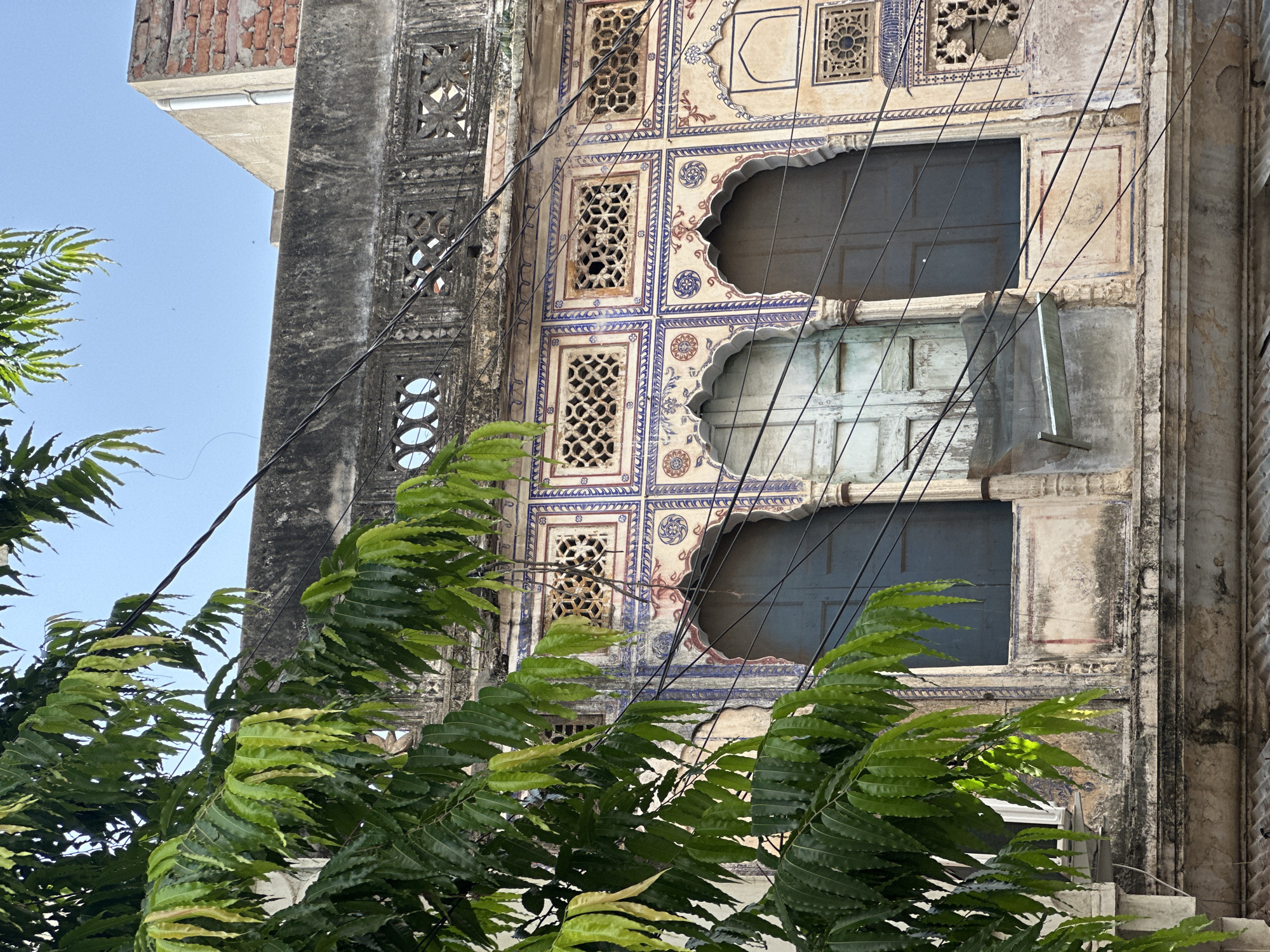 This image depicts the exterior of a traditional building in Pushkar, India, characterized by its ornate architecture. The facade features arched windows with dark wooden shutters, framed by intricate decorative patterns in blue and red. Above the windows, there are several smaller square openings with latticework, possibly serving as ventilation or decorative elements. The building appears aged, with weathered plaster and some signs of decay, but retains its historical charm. Lush green foliage from a tree in the foreground partially obscures the view of the building, adding a natural element to the urban scene. Overhead, electrical wires crisscross the frame, indicative of the modern infrastructure overlaid on the historic setting. The sky above is a clear blue. The scene captures a blend of old-world architecture and everyday life in an Indian city.