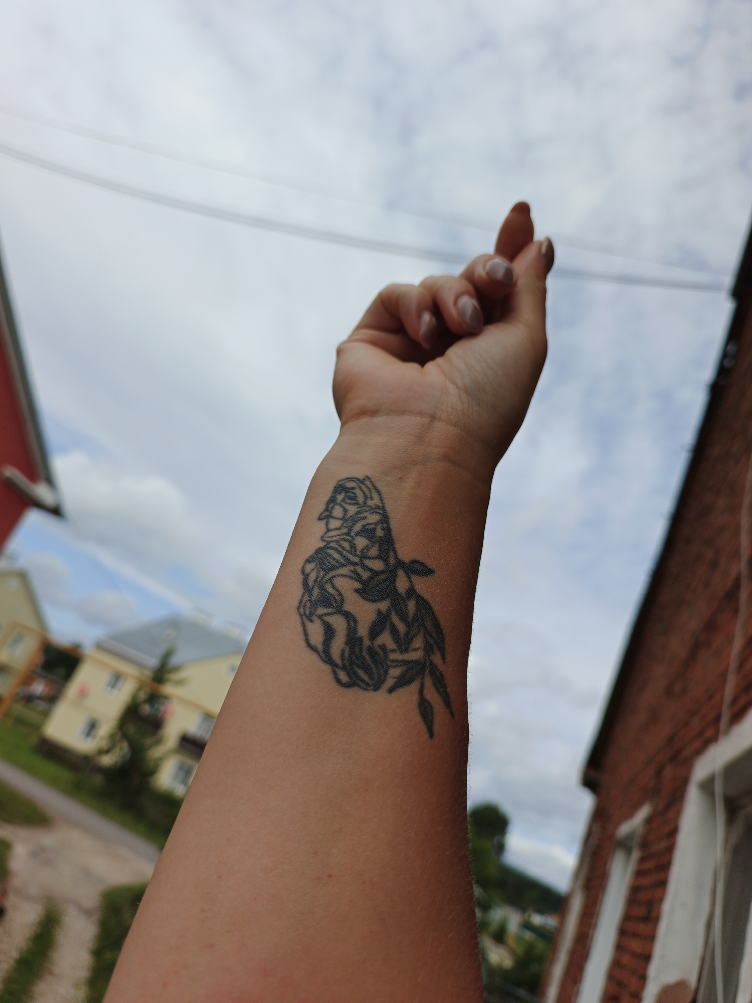 A close-up shot focuses on a person's arm, displaying a black line-art tattoo on the forearm. The tattoo depicts a stylized face or bust interwoven with foliage, rendered in a minimalist design. The arm is angled upwards against a backdrop of a cloudy sky. In the mid-ground, blurred residential buildings and greenery are visible, suggesting an outdoor urban or suburban setting. The lower portion of a brick building frames the right side of the image. This scene is located in Medvedyovka, Russia.