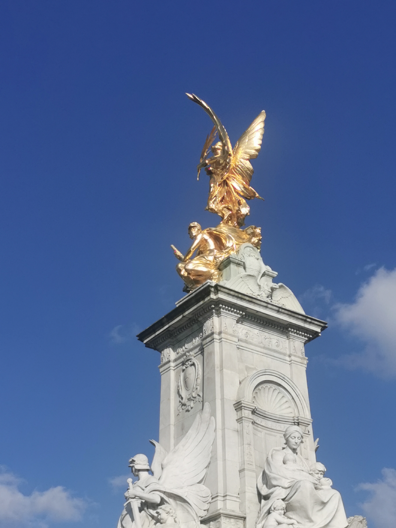The image depicts the Victoria Memorial, located in front of Buckingham Palace in London, United Kingdom. The memorial features a large, multi-tiered white marble base adorned with various sculptures and carvings. At the apex is a gilded bronze statue of Victory, a winged female figure, standing atop a globe. Below Victory are figures in gilded bronze, depicting courage and constancy, seated on the base. Additional white marble sculptures are positioned around the base, including representations of maritime power, manufacture, peace, progress, agriculture, and an image of Motherhood. The sky is a vivid blue with scattered white clouds.
