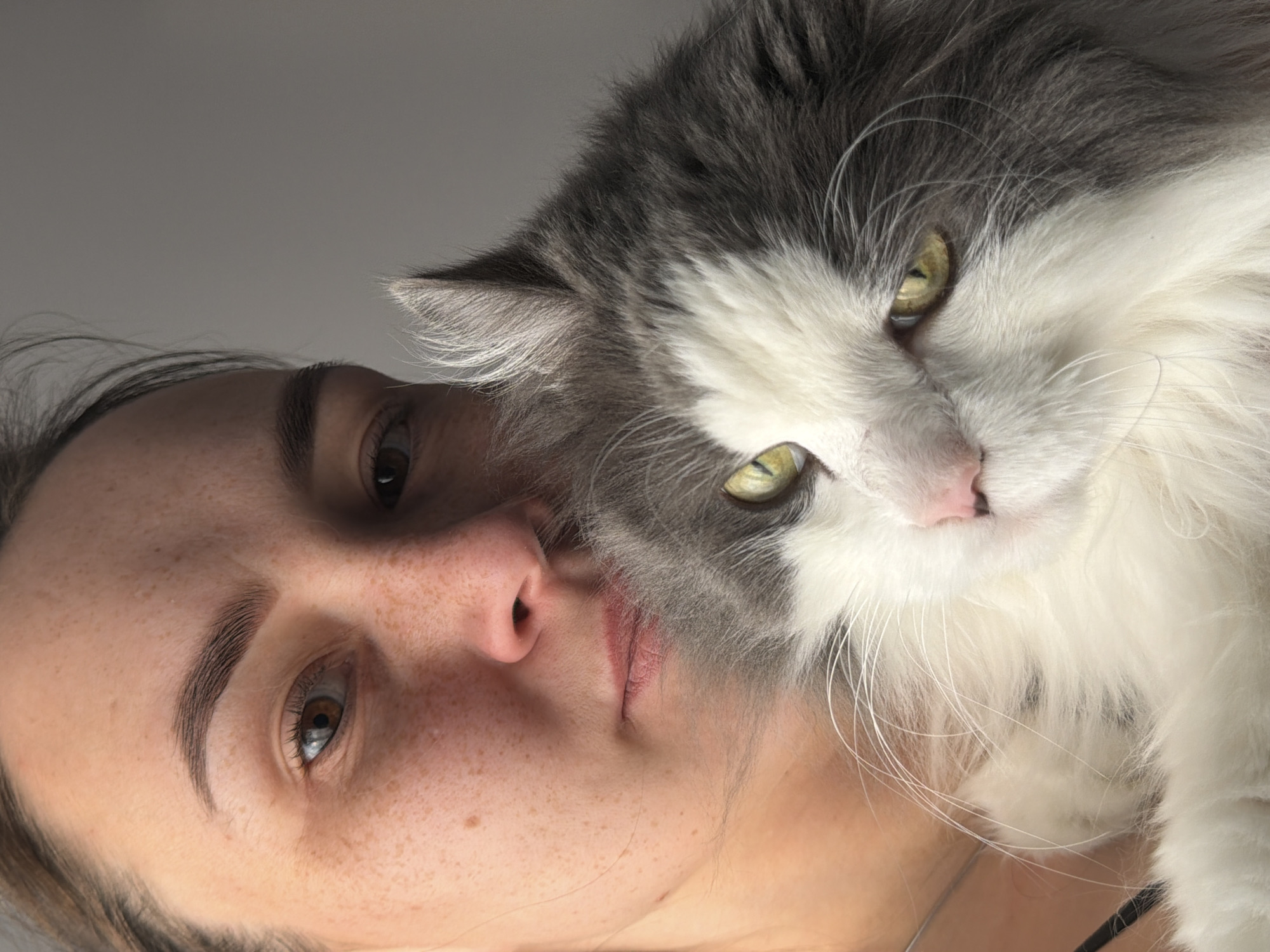 A human female with freckles is closely juxtaposed with a long-haired, bicolor cat (gray and white). Both face the camera. The setting or location in Chaikovsky, Russia, is not discernible from the visual content.