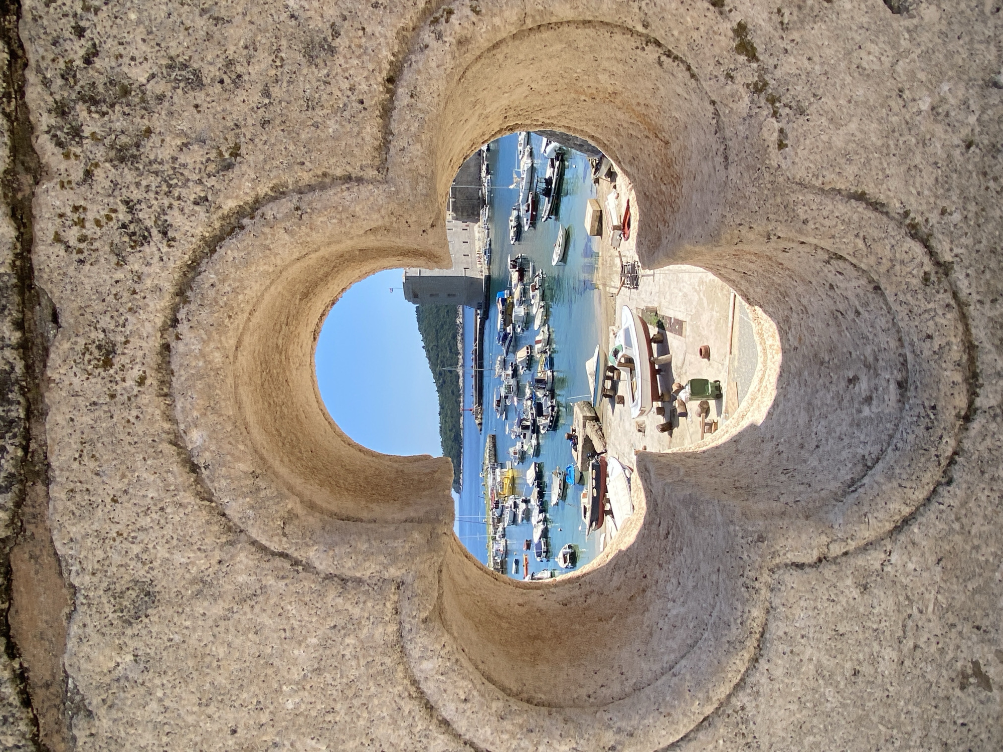 This image, viewed through a quatrefoil-shaped opening in weathered stone, depicts a picturesque harbor in Dubrovnik, Croatia. The opening frames a vibrant scene of a bustling marina under a clear blue sky. Numerous boats of various sizes, from small fishing vessels to larger yachts, are moored in the turquoise water. Some boats are docked along the stone quayside, while others float freely.
In the background, a prominent stone tower, part of the city's historic fortifications, rises majestically above the harbor. The tower's architecture suggests a historical significance, likely a defensive structure common in Dubrovnik. Lush green hills dotted with vegetation form the backdrop, further enhancing the scenic beauty of the location. The stone detailing of the opening suggests it is part of an ancient architectural element, possibly a window or decorative feature from a historical building within the city. The scene evokes a sense of timeless charm and the maritime heritage of Dubrovnik.
