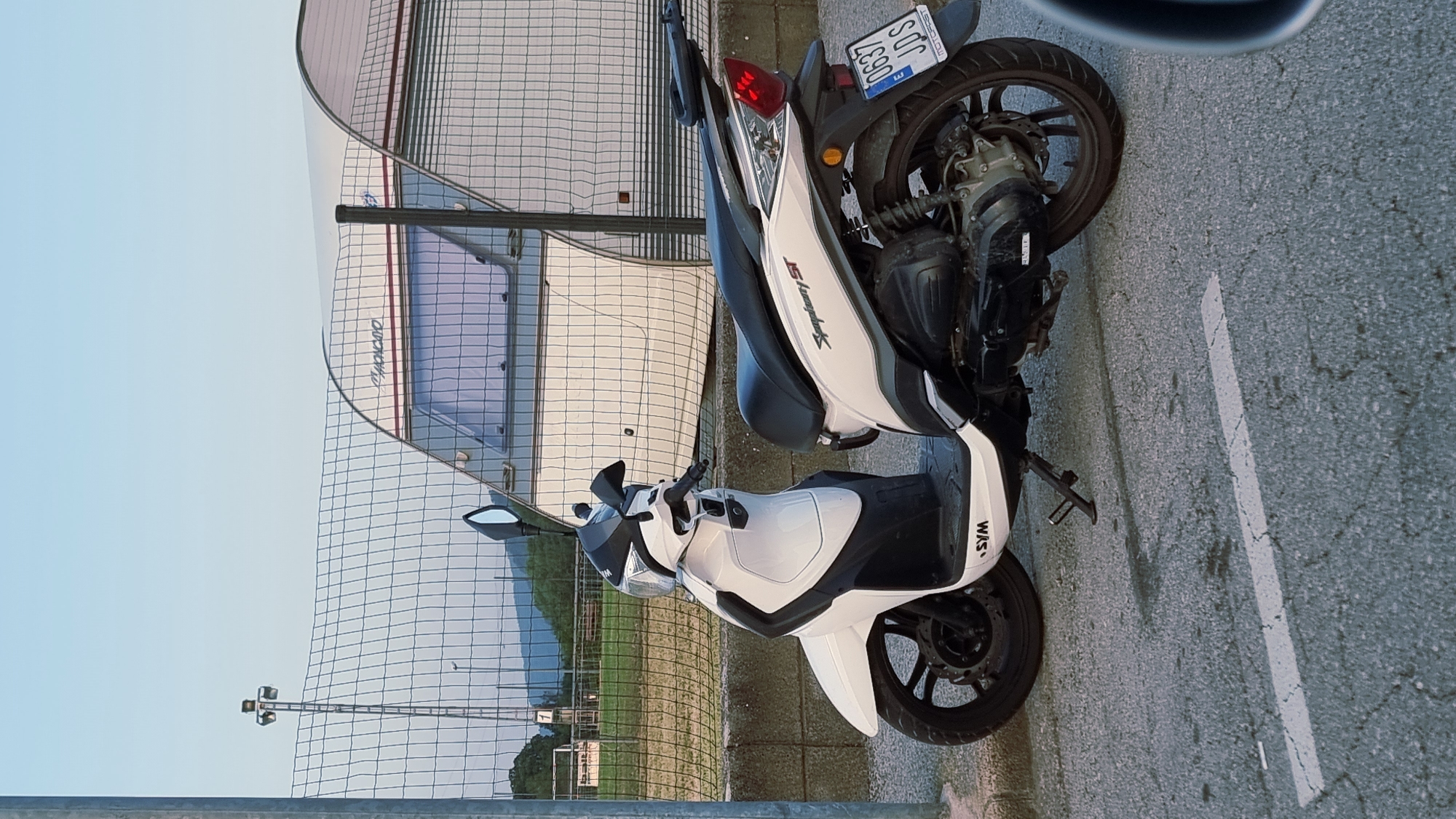 A white and black Sym Symphony ST scooter is parked on a cracked asphalt surface with a white stripe. The scooter is facing right, with its kickstand down. It has a black seat and black wheels. A license plate that reads "0637 JDS" is visible on the rear. Behind the scooter is a metal fence, a concrete wall, and a white caravan. In the background, there are green fields, trees, and mountains under a clear blue sky. A tall metal pole with lights is also visible to the left of the caravan. The scene appears to be in a rural or semi-rural area near La Canya, Spain.