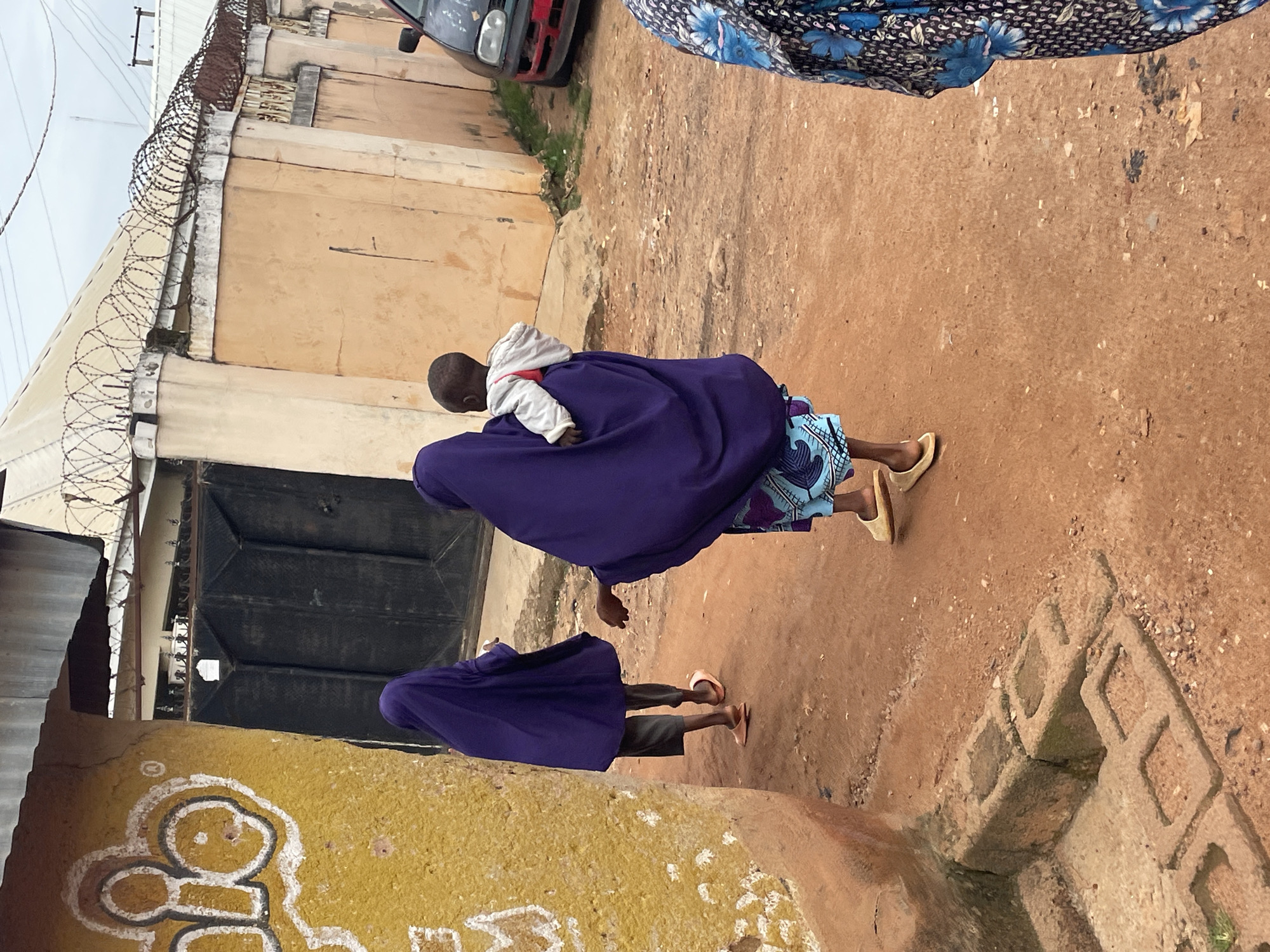 Two people, one carrying a baby, walk along a dirt path in front of a yellow building with white painted symbols. The person in front is wearing a purple hijab and grey pants, and the person behind is also wearing a purple hijab and a patterned blue and white skirt. They are walking away from the camera towards a large black gate. In the background, a wall with barbed wire runs along the top. To the right, a black car with a red bumper is parked. This scene appears to be in an urban environment, possibly the city of Jos, Nigeria.