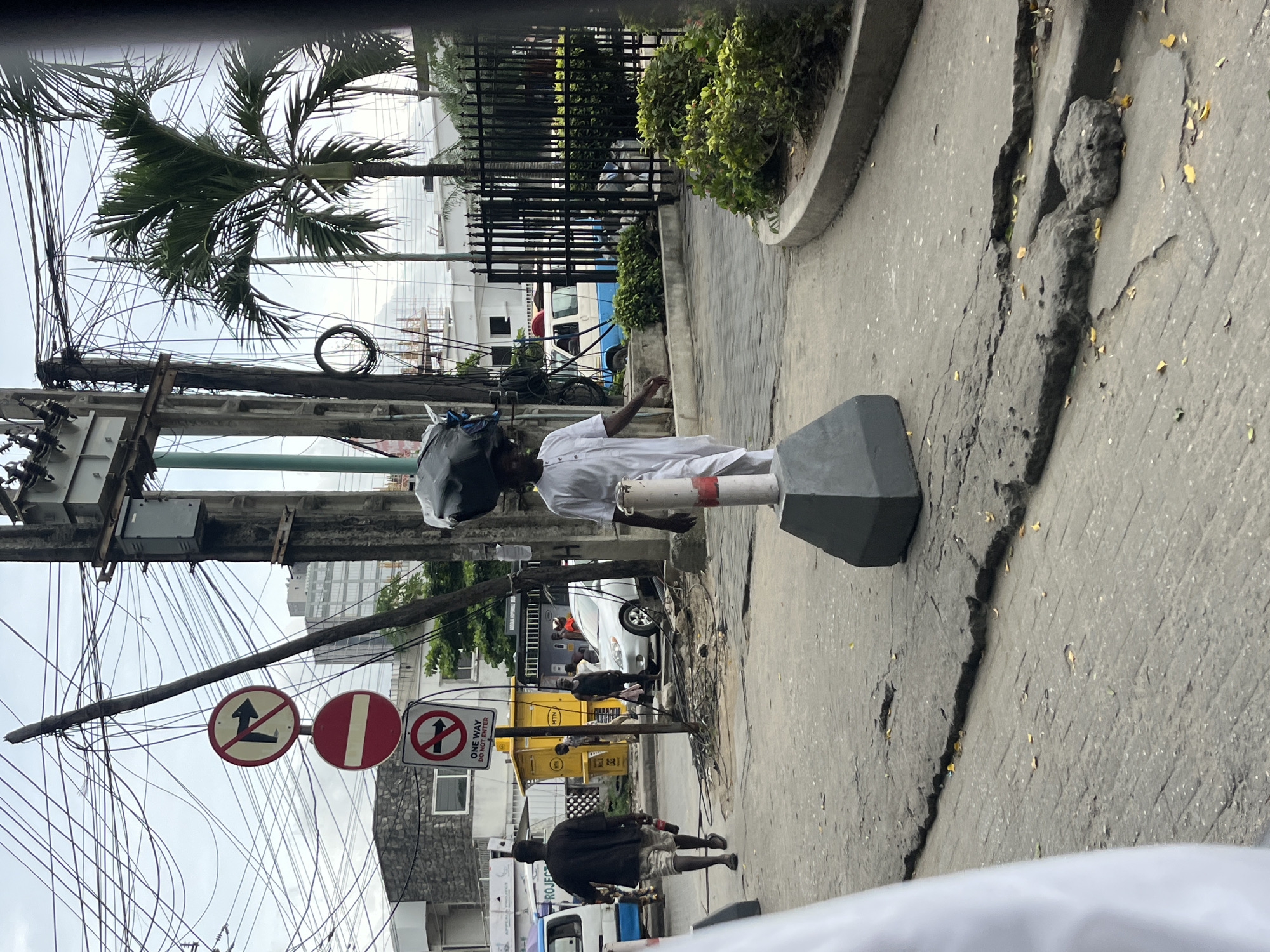 The image depicts a street scene in Itirin, Nigeria. A man in a white shirt and carrying a black bag on his head stands next to a grey street bollard on a cracked pavement. Overhead, a tangle of power lines is attached to a utility pole. Traffic signs indicating restrictions are visible near the pole. In the background, there are buildings, parked vehicles, and other pedestrians. A palm tree is visible against the sky.