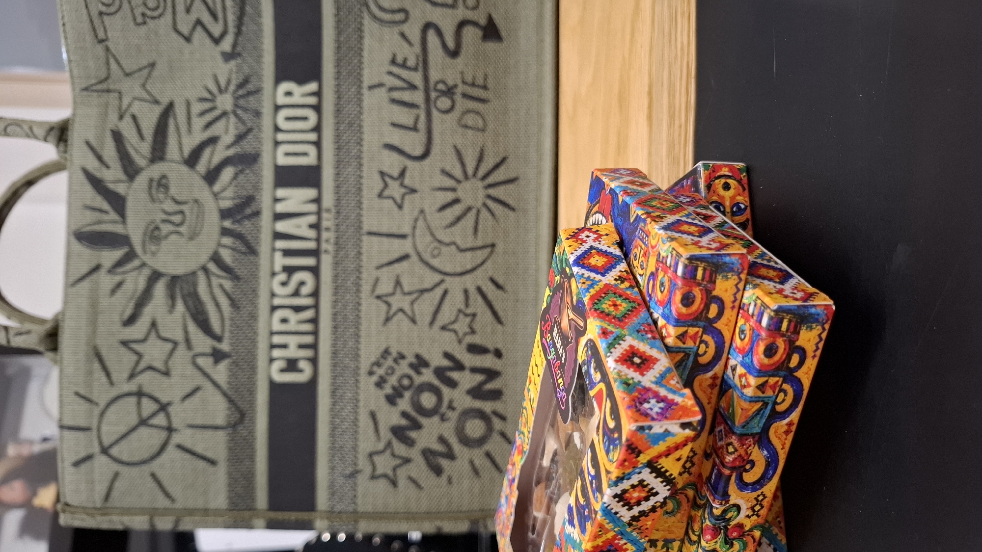 The image shows a close-up of stacked boxes with colorful, geometric patterns. In the background, a green tote bag with "CHRISTIAN DIOR" written in large letters is visible. The bag is decorated with various drawings, including a sun with a face, stars, a peace sign, and the words "LIVE OR DIE". The scene appears to be indoors, possibly on a table. There are no people or animals in the image. The photograph does not contain any information that would allow it to be located in Plovdiv, Bulgaria or any other specific city.