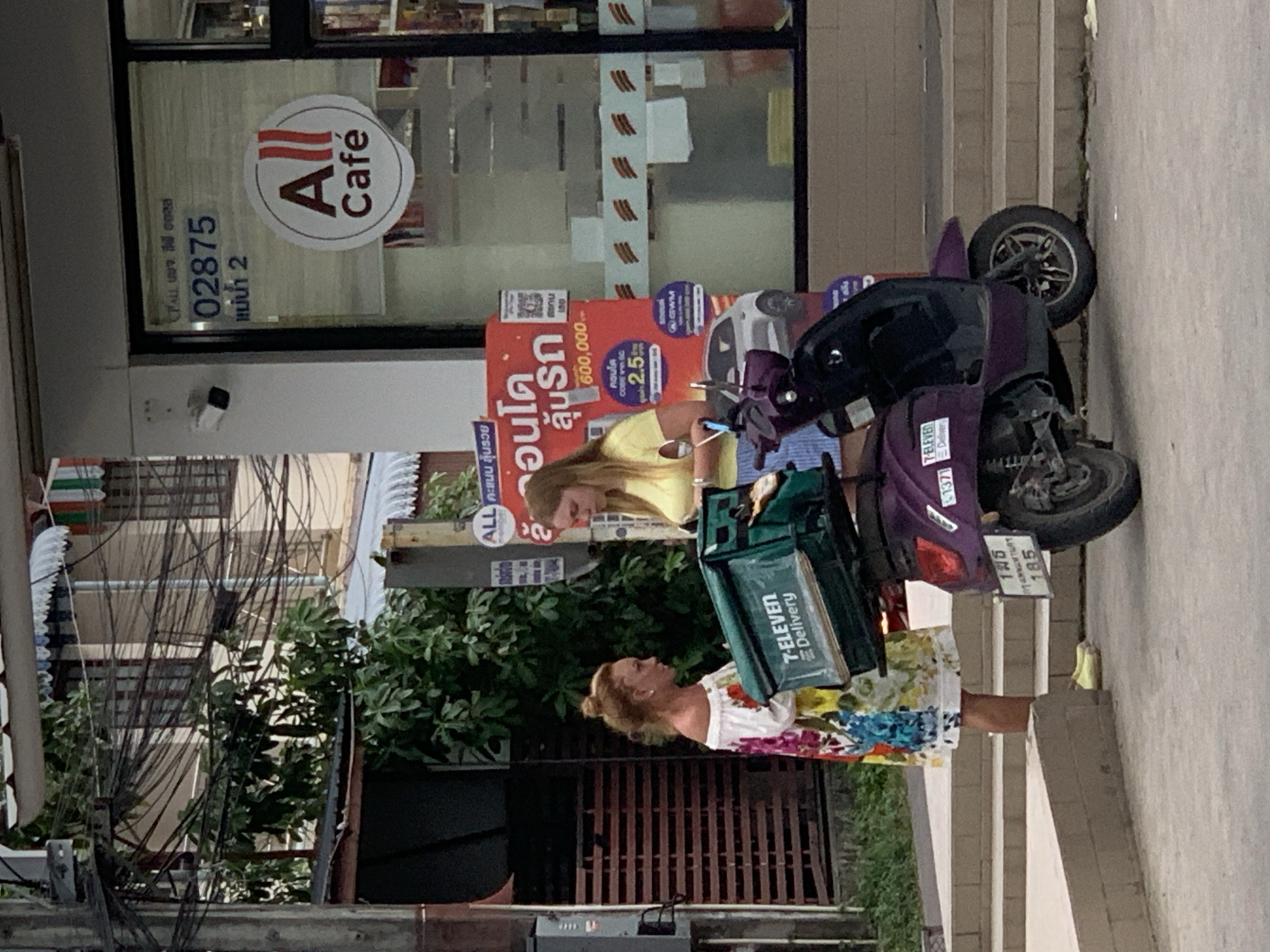 The image captures a street scene in the city of Ko Samui District, Thailand. In the foreground, a purple motor scooter is parked, equipped with a green 7-Eleven delivery box on the back. Two women are standing next to the scooter. One woman, on the left, wears a colorful off-the-shoulder dress and has her hair in a bun. She is facing the other woman and appears to be interacting with her. The second woman, on the right, wears a yellow t-shirt and blue striped shorts. She is looking down and appears to be engaged in conversation with the first woman. Behind the individuals and the scooter, a building with a glass storefront is visible, featuring a sign that reads "ALL Café" and other text. The building also has a security camera mounted on its exterior. The background shows more of the urban environment with electrical wires and trees.