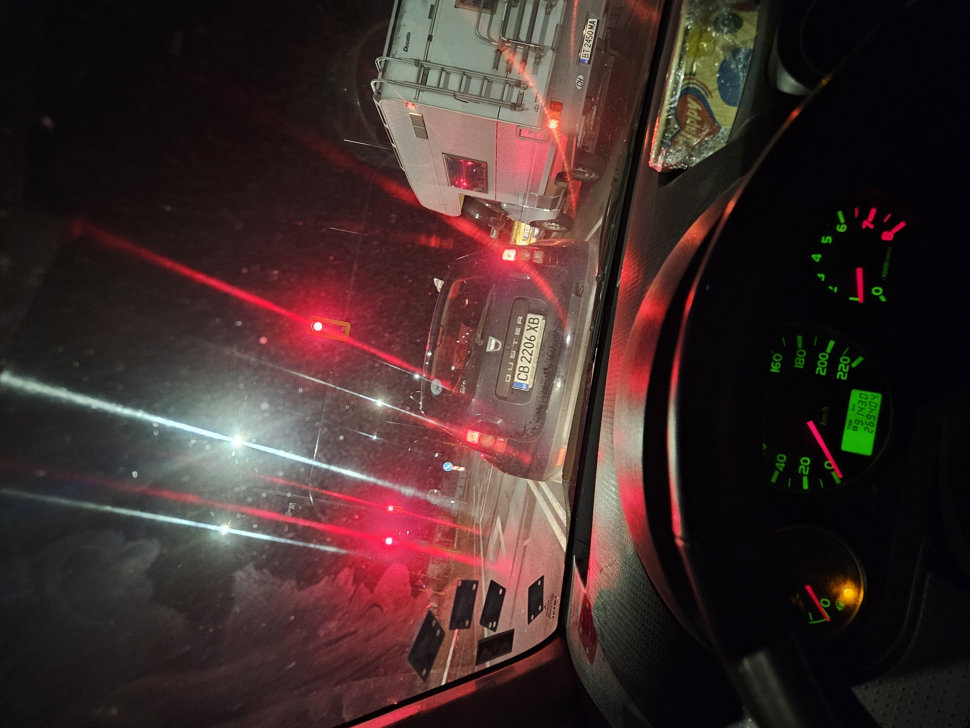 The image is taken from the interior of a car at night, looking forward through the windshield. The car is in traffic, indicated by the red brake lights and the presence of other vehicles. A Dacia Duster is in front and to the left, with a license plate that reads "CB 2206 XB". To the right, a white camper van is visible, with a license plate that reads "BT 2450 MA". Traffic lights are illuminated red, casting a red glow on the scene. The dashboard of the car is visible in the foreground, with illuminated gauges showing speed and engine RPM. There are multiple black rectangular stickers on the windshield. The overall environment suggests a congested urban street at night.
