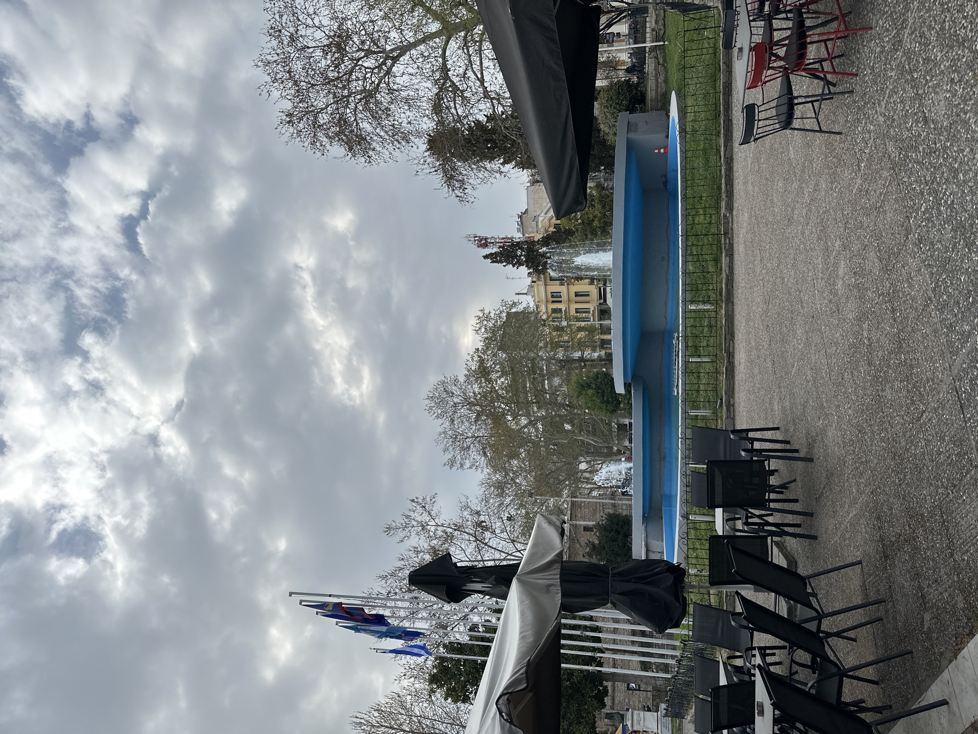 The image depicts a scene in the city of Serres, Greece, likely in a public square or park. A blue fountain with a metal fence is prominently featured. Several chairs are arranged near the fountain, some of which are black metal chairs typical of outdoor seating, while others are red and black. The foreground includes covered parasols, suggesting a cafe or outdoor eating area. Flagpoles with flags are visible. In the background, buildings typical of a European city can be seen through bare trees. The sky is overcast with heavy clouds. The ground appears to be a paved area. A water fountain is visible in the background.