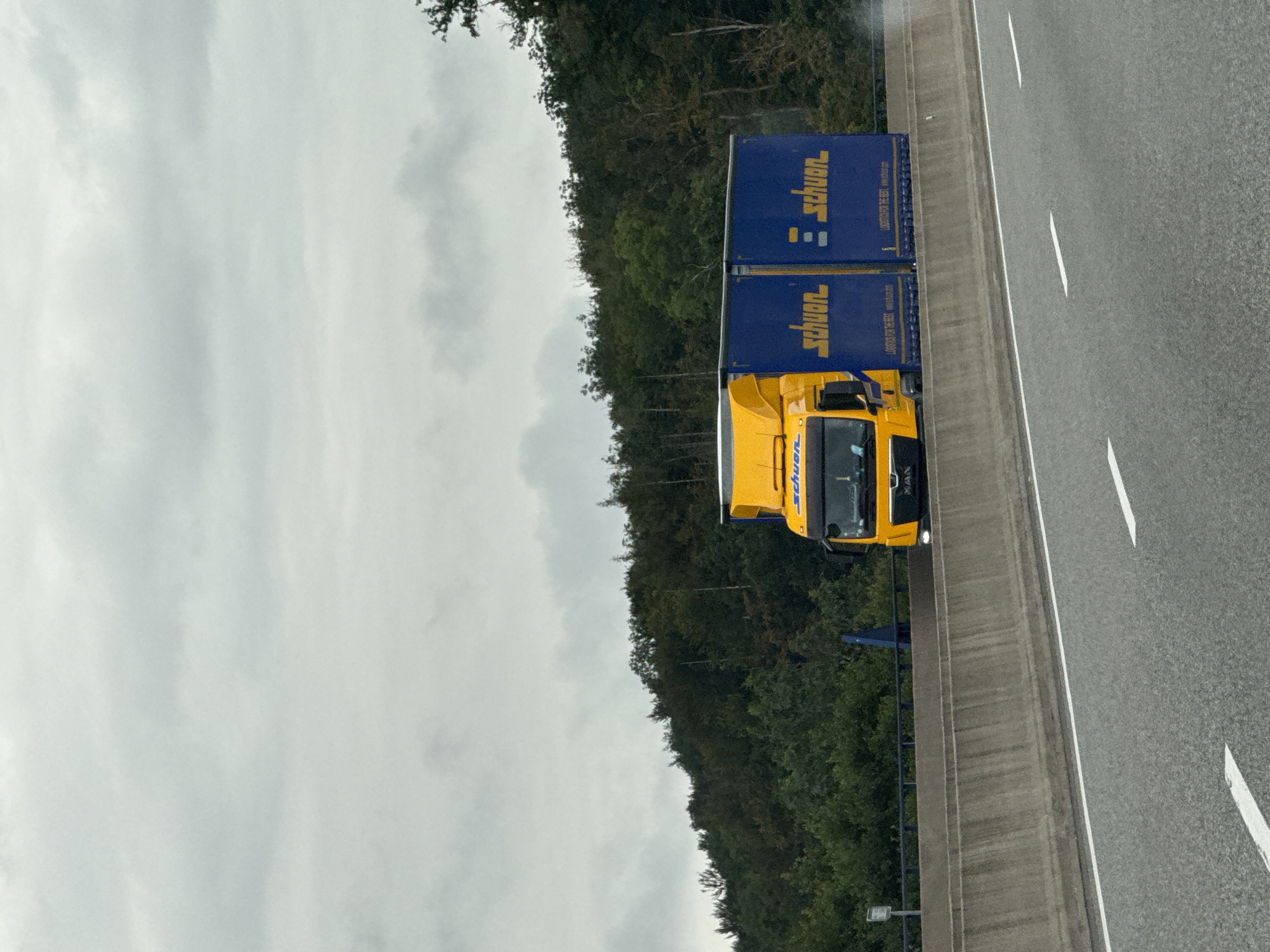 An outdoor, eye-level shot captures a yellow and blue semi-trailer truck driving on a highway. The truck is branded with "schuen" in yellow lettering on both its cab and trailer. The trailer is blue and has additional yellow text. The truck is positioned on the right side of the frame, with the road and a concrete barrier in the foreground. Beyond the barrier, a dense, green forest with tall trees lines a hillside. The sky is overcast and grey. The image is likely taken in the vicinity of Bad Lauterberg im Harz, Germany, given the context.