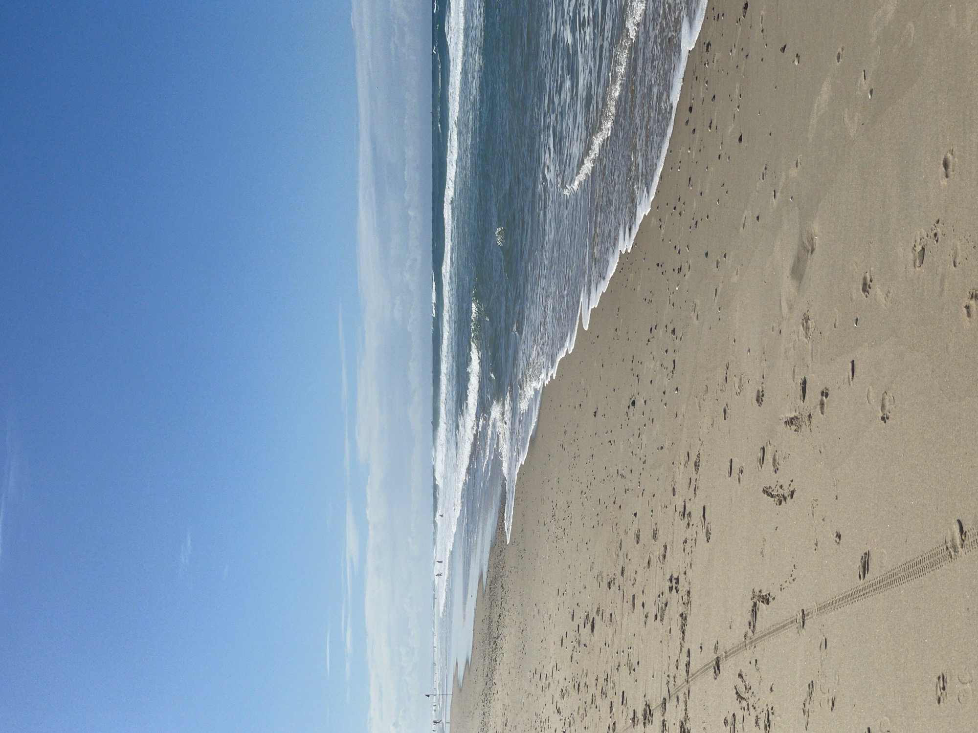 The image depicts a wide, sandy beach with gentle waves rolling in from the ocean under a clear blue sky with scattered clouds. The foreground shows a textured sand surface with numerous footprints and a distinct tire track pattern from a vehicle. In the middle ground, the ocean is a deep blue-green, with white foam from the breaking waves. A few distant figures, appearing as small silhouettes, are visible along the shoreline and in the water. The horizon line separates the ocean from the sky, with a thin line of clouds in the distance. The overall impression is a serene, natural beach scene.
While the image captures a typical beach environment, the specific location within Surfers Paradise, Australia, cannot be determined from the visual information alone. Surfers Paradise is known for its wide, golden beaches and the consistent surf, which aligns with the scene depicted. However, without any identifying landmarks or signage, pinpointing the exact spot within Surfers Paradise is not possible based on the provided image.