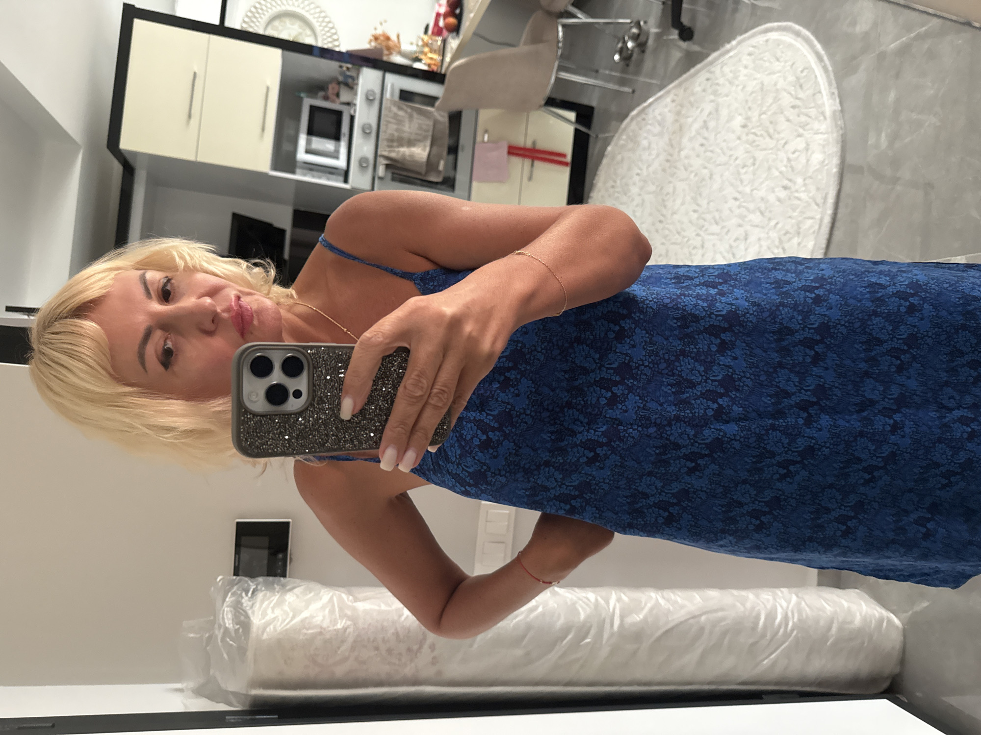 A blonde woman stands in what appears to be a kitchen or studio apartment in Alanya, Turkey, taking a mirror selfie. She is wearing a blue floral print dress with spaghetti straps and a gold necklace. Her hair is styled in loose waves, and she is wearing makeup. She holds a smartphone with a sparkly case in her right hand, and her left hand is on her hip. In the background, a large roll of what looks like carpet or protective sheeting is visible against a white wall. The kitchen area features cream-colored cabinets, a microwave, and a stainless steel oven. A dining chair and a white oval rug are also in the frame. The overall lighting suggests an indoor setting during the daytime.