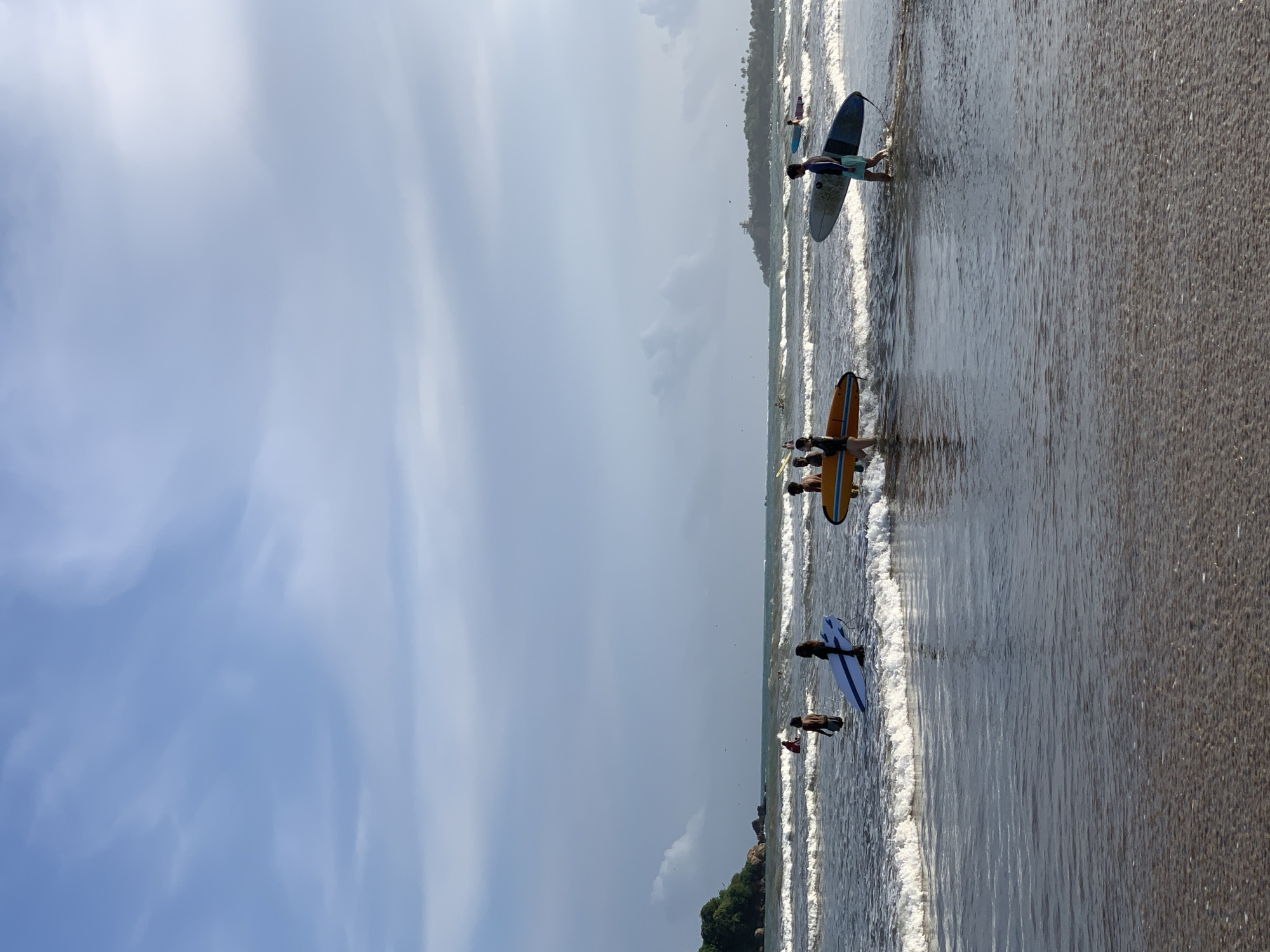 The image depicts a beach in Weligama, Sri Lanka. The scene includes several people with surfboards in the water. They appear to be walking and standing in the shallow water near the shoreline. The sky is overcast with a mix of blue and gray clouds. The sand is dark, and gentle waves are visible. Some trees and vegetation are visible along a shoreline in the distance.
