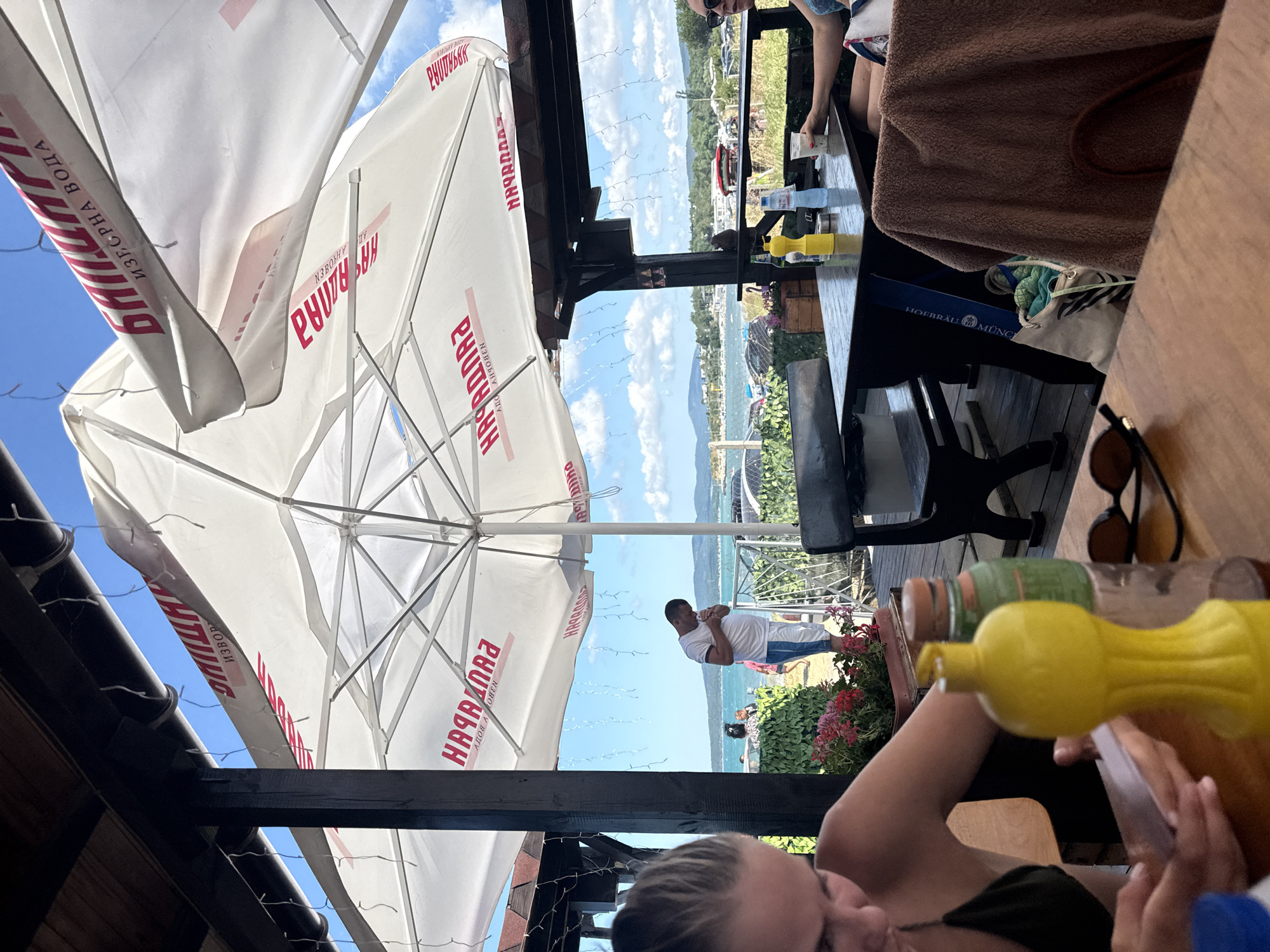 This is a photograph taken outdoors, likely at a waterfront establishment, possibly a restaurant or beach bar, in Burgas, Bulgaria. The scene is oriented vertically. In the foreground, the upper bodies and faces of two people are visible, one partially obscured by a brown towel draped over a chair. They appear to be seated at a wooden table, where several bottles and condiments are placed, including a yellow squeeze bottle and a green bottle.
Dominating the upper portion of the frame are large white umbrellas with red lettering, providing shade. The text on the umbrellas, visible in Cyrillic script, includes "ИЗВОР" (Izvor, meaning source) and "НАГРАДА" (Nagrada, meaning prize or award), along with "ИЗВОРНА ВОДА" (Izvorna Voda, meaning spring water). One umbrella has additional text that appears to be "ХОФБРАУ МЮНХЕН" (Hofbräu München), suggesting a possible connection to the German beer brand.
In the background, beyond a wooden railing and some greenery, a body of water is visible, likely the Black Sea, with boats and distant hills. A man in a white shirt and blue shorts stands near the water, appearing to hold something to his mouth. The sky is blue with scattered white clouds, indicating a bright, sunny day, likely midday. The overall mood appears relaxed and leisurely, consistent with a vacation or leisure setting.