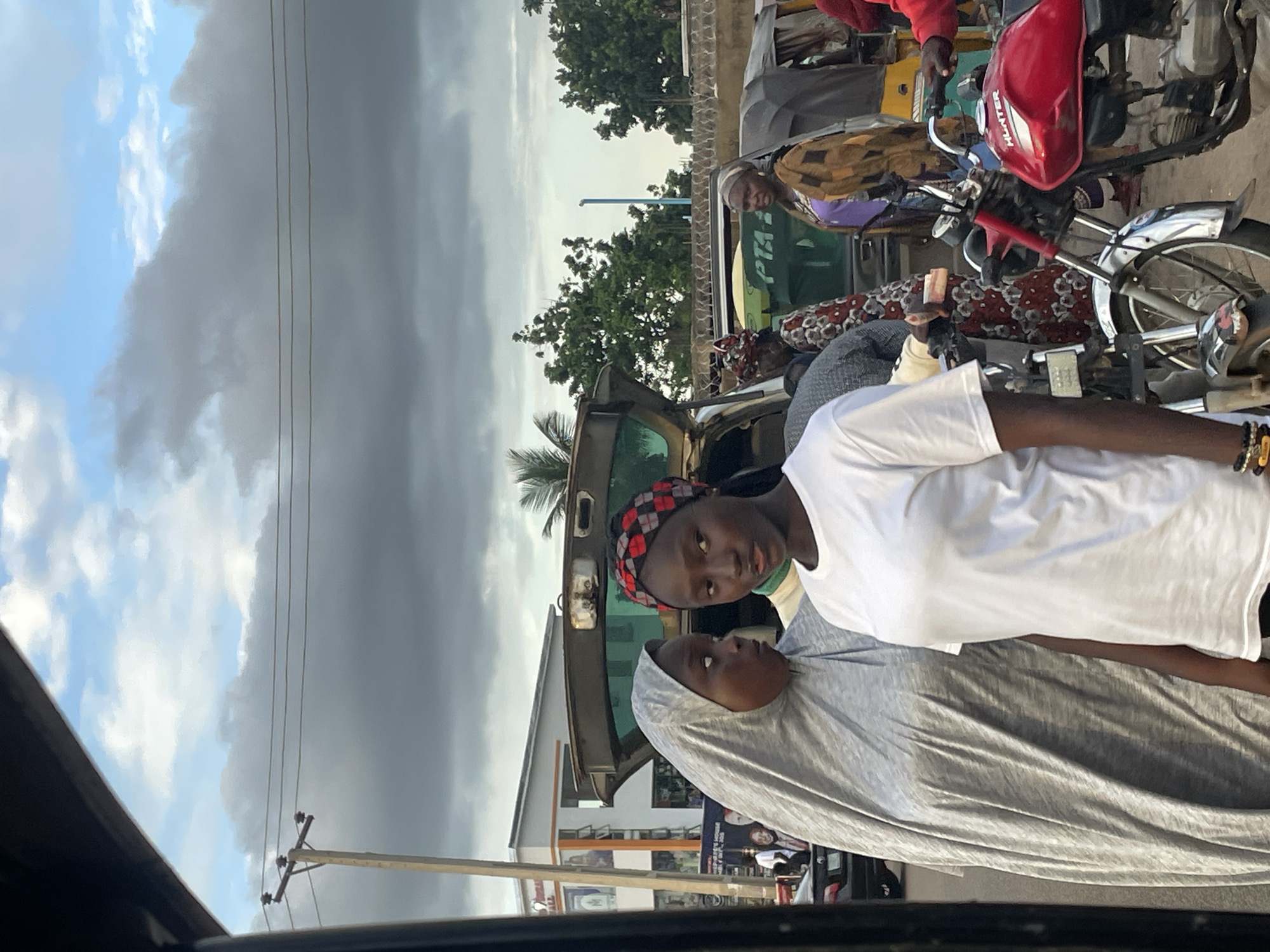 The image shows a street scene in Jos, Nigeria, with several people and vehicles. Two young women are prominently featured in the foreground. One is wearing a grey hijab and a white shirt, while the other has a patterned headscarf and a white t-shirt. Behind them, an open car trunk reveals people and some luggage. Various other individuals are visible, some in traditional attire, standing near or interacting with a motorcycle and a three-wheeled vehicle, likely a tuk-tuk or auto-rickshaw. In the background, there are buildings, trees, a utility pole with wires, and a cloudy sky. The overall impression is of a bustling urban environment, possibly a transportation hub or a marketplace.
