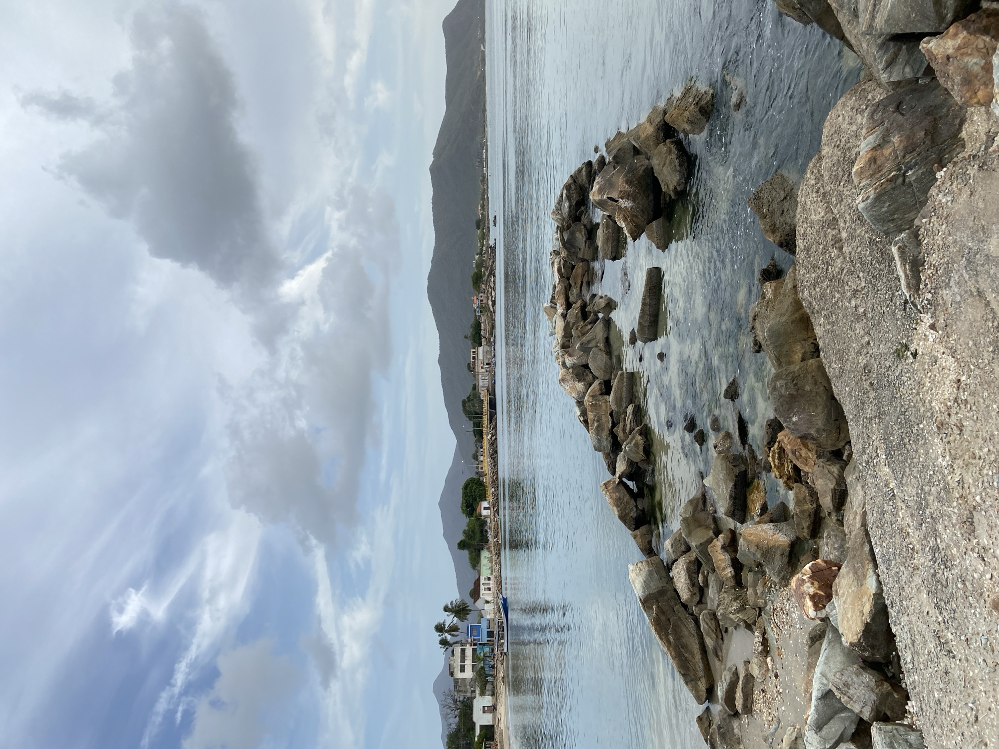 The image depicts a serene coastal scene in Juan Griego, Venezuela. In the foreground, a rocky embankment leads into calm, clear water. A breakwater made of large stones juts out into the sea, with smaller rocks and pebbles scattered along the shore. In the middle ground, a small village lines the coastline, featuring a mix of residential buildings and some palm trees. A blue boat is moored in the shallow water near the shore. The background is dominated by rolling hills and mountains, under a sky filled with a mix of scattered clouds and wispy cirrus formations. The overall impression is one of a tranquil, picturesque coastal town.