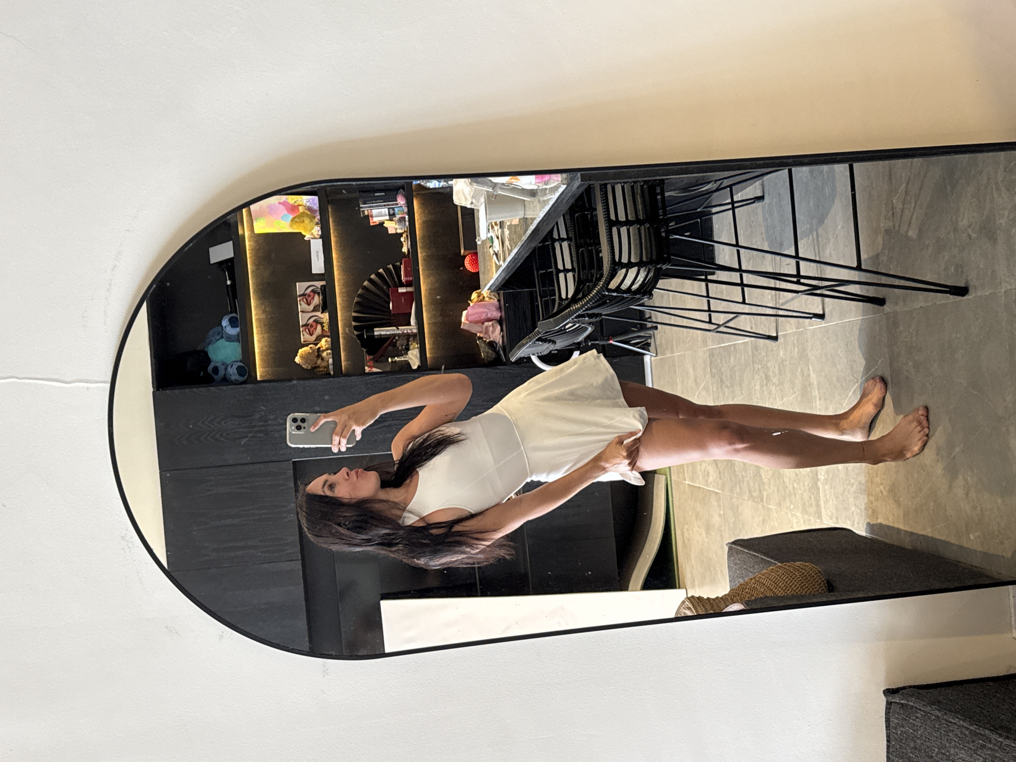 A full-body, mirrored selfie captures a woman in a white, form-fitting athletic dress with a flared skirt. She stands in what appears to be a modern, indoor space, likely a residence, in North Kuta, Indonesia. The woman is facing a large, arched mirror, holding a silver smartphone in her right hand and taking the picture. Her left hand is slightly lifting the hem of her dress. She has long, dark hair and is looking at her reflection with her lips pursed.
Behind her, visible in the mirror, is a sleek, dark wood shelving unit filled with various items, including what appear to be decorative objects, boxes, and possibly some small stuffed animals. The shelving unit has integrated lighting, casting a warm glow on the displayed items. To the right of the shelves, a black marble-topped counter with bar stools is visible. The stools have black metal frames and woven seats. The floor is tiled with large, gray square tiles. In the foreground of the mirrored reflection, on the left side, a portion of a dark gray upholstered bench is visible, with a woven straw bag resting on it. The overall environment suggests a contemporary living space.