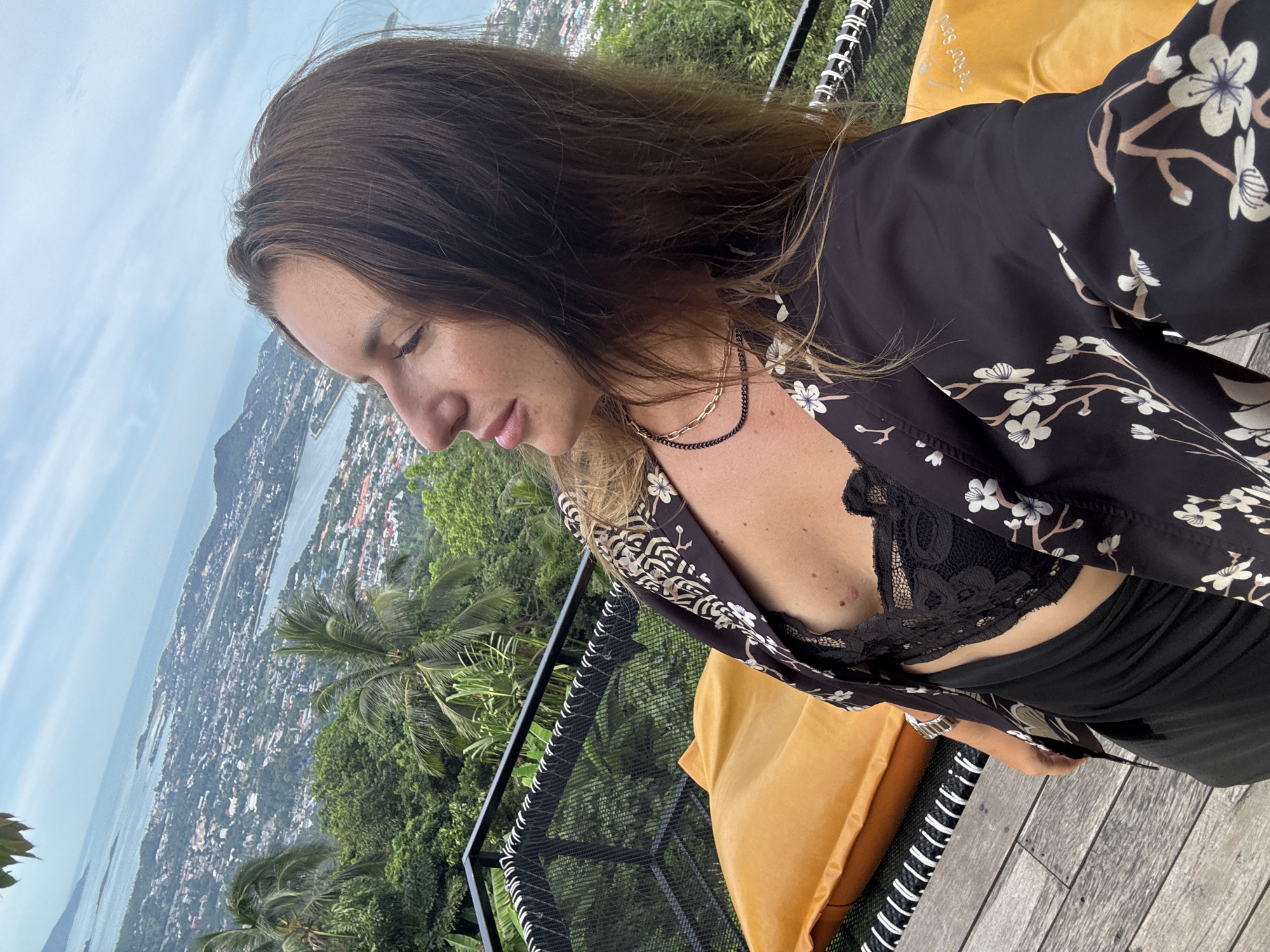 A woman with long brown hair is standing on a wooden deck overlooking a city and ocean. She is wearing a black lace bra, a black skirt, and an open black kimono with a floral pattern. She is looking to her right, with her mouth slightly open. There is a yellow cushion behind her. The deck has a black net railing. The city is spread out below, with buildings and a body of water. There are palm trees and other greenery in the foreground. The sky is cloudy.