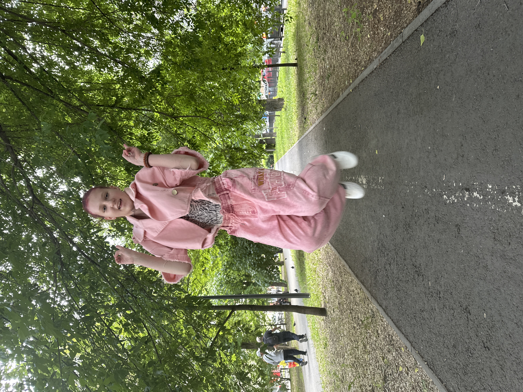 A young person with a shaved head stands on a paved path, likely in a park, in Saratov, Russia. The person is wearing a pink jacket, pink pants, and white shoes. They are facing the camera and pointing upward with both hands. There are trees on either side of the path. In the background, other people can be seen walking on the path.