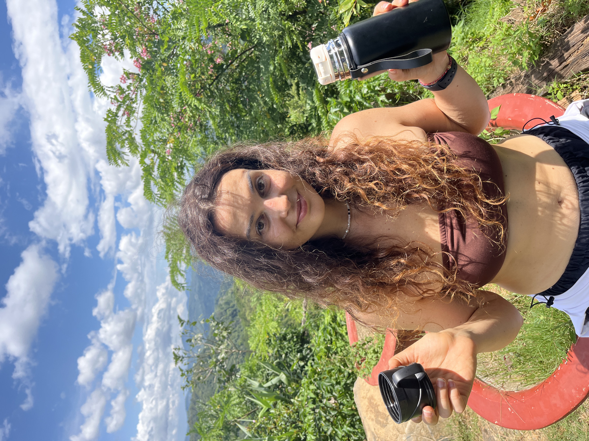 A woman stands outdoors with a lush green landscape and cloudy blue sky behind her. She is holding a black thermos in her right hand and a black lid in her left hand. She is wearing a brown strapless top and white and black shorts. The location appears to be a scenic overlook in Son Dien, Vietnam, with rolling hills and vegetation visible in the background.