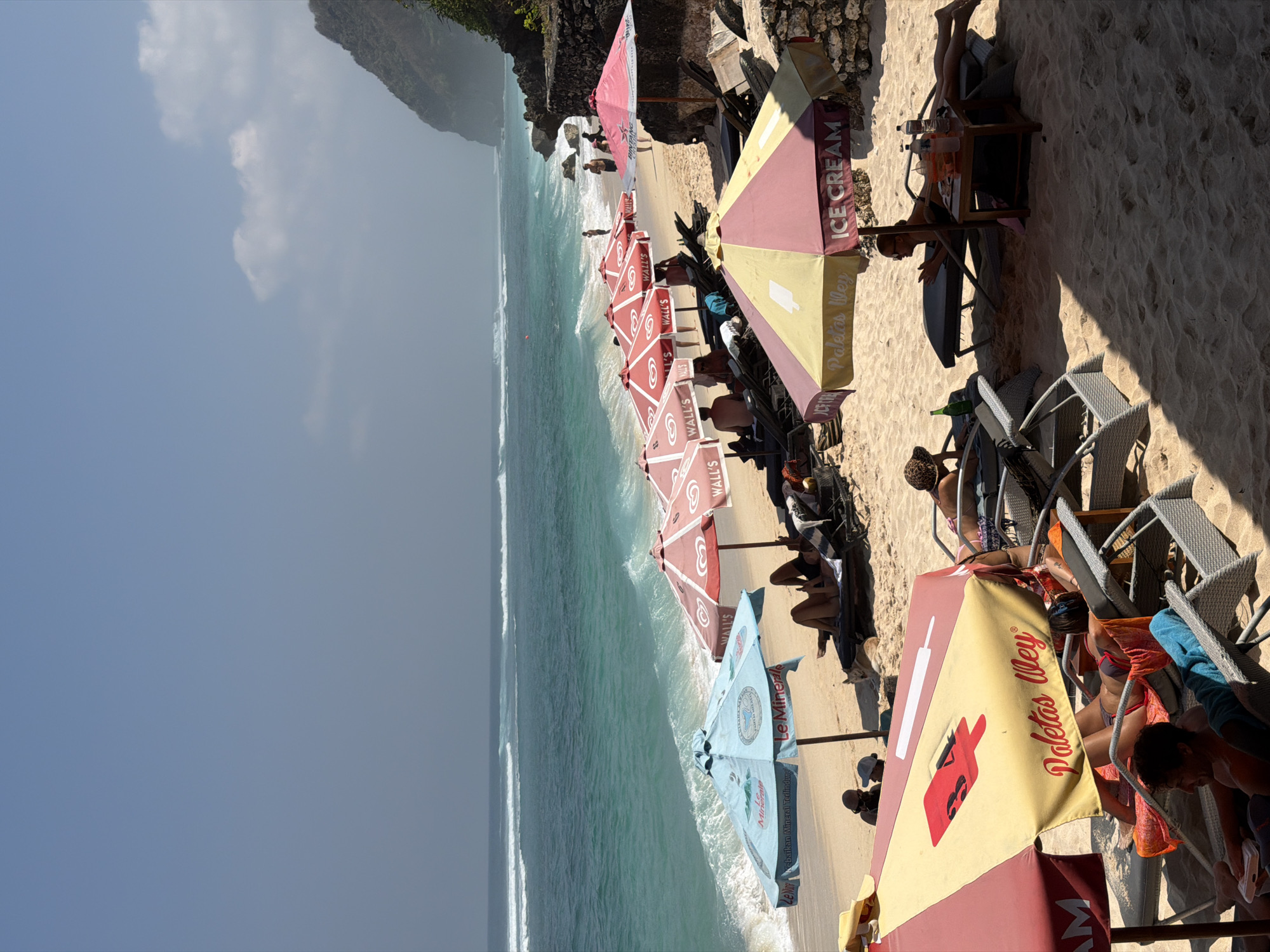 An eye-level, medium shot captures a sun-drenched beach scene with several umbrellas and lounge chairs on a sandy shore. The ocean, a vibrant turquoise, features gentle waves rolling in under a hazy blue sky with scattered clouds. In the background, a rocky cliff face rises from the right side of the frame.
Underneath a light blue umbrella, partially visible is a person wearing a dark top. To the right, beneath a red and white umbrella advertising "WALL'S ICE CREAM," two people recline on lounge chairs. One individual, wearing a black bikini, faces away from the camera, while another person, whose head is not visible, is also seated.
Further to the right, a pink umbrella also advertises "WALL'S ICE CREAM." In front of it, a person lies on a lounge chair wearing a black swimsuit and a brown hat. Beside them is a small wooden table with several bottles and cups. Another person is visible further back, sitting on a lounge chair.
In the foreground, under a large yellow and red umbrella featuring a red popsicle graphic and the text "Paletas Wey ICE CREAM," two people are seated on lounge chairs. The person on the left, with dark hair, wears a patterned hat and sunglasses and is looking downwards. The person on the right, wearing a bikini, is reclined and looking towards the ocean. Another lounge chair in the very foreground is occupied by a person with curly hair, wearing a blue shirt and holding a white object.
The sand is golden and appears well-trodden, with shadows cast by the umbrellas indicating a bright, sunny day. The overall atmosphere is one of relaxation and leisure.