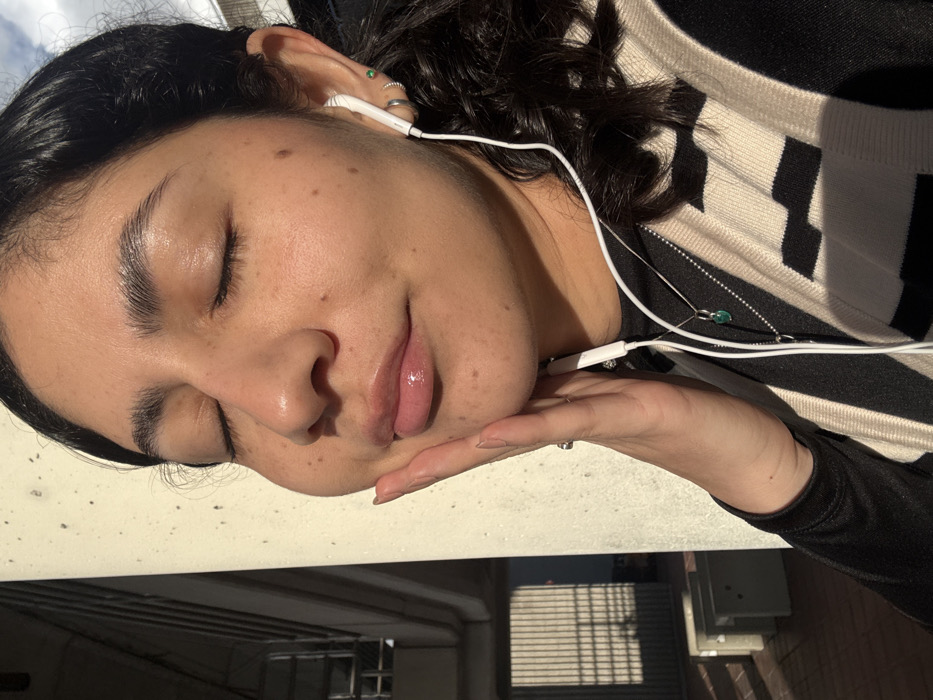 A close-up outdoor photograph depicts a female subject located in Hialeah, United States of America. The subject has medium skin tone, dark wavy hair, and visible freckles on her face. Her eyes are closed, and her lips are slightly parted, exhibiting a glossy appearance. Her left hand is positioned with fingers resting gently on her left jawline and cheek. She is wearing white wired earphones in both ears. The visible right ear features a green stud piercing, two silver hoop earrings, and an additional silver ring piercing. Around her neck, two silver chain necklaces are present; one has a small green heart-shaped pendant. She is dressed in a black long-sleeved top overlaid with a beige and black vertically striped vest. A silver ring is visible on her left ring finger. Strong, direct sunlight illuminates the subject's face from the right, casting a bright sheen on her skin and hair. The immediate background features a large, light-colored concrete pillar to the subject's left. Further in the background, out-of-focus urban infrastructure, including metallic vertical elements and a concrete surface with distinct shadows, is discernible, indicative of a structure like an underpass or parking garage. The lighting suggests a low sun angle.