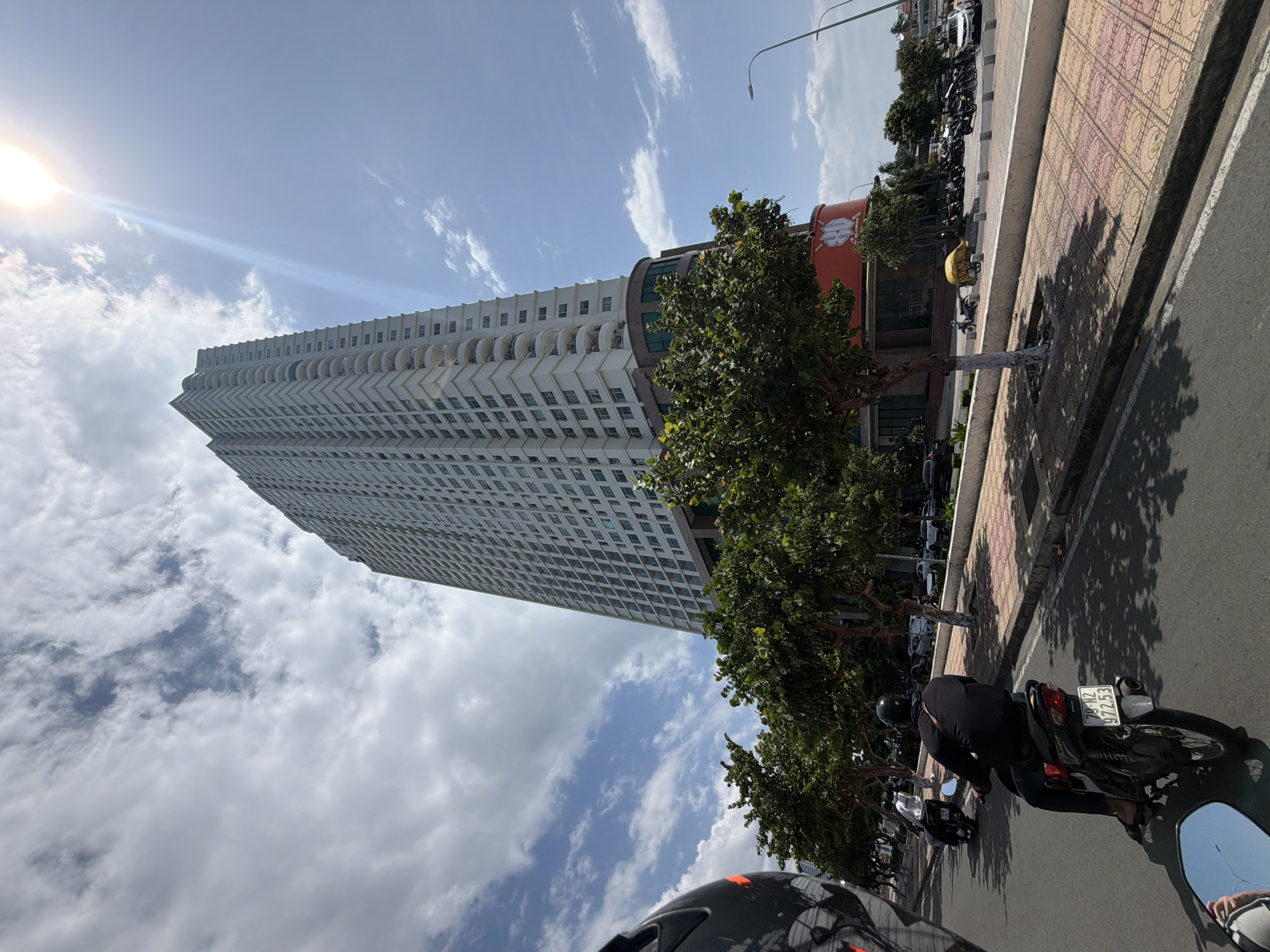 The media file, originally rotated ninety degrees counter-clockwise, captures a sunlit urban street scene in Bac Nha Trang Ward, Vietnam. A prominent, modern, multi-story white building with a distinctive facade of numerous balconies rises sharply on the left side of the frame, contrasting with a bright blue sky adorned with scattered white clouds. The sun is visible in the upper left, producing a strong lens flare.
On the right, a street runs parallel to a wide sidewalk paved with patterned tiles. Green trees line the sidewalk, casting sharp shadows on the ground, indicative of a clear, sunny day. In the immediate foreground, from the perspective of a motorcyclist, the back of another person wearing a black helmet and black attire is visible, seated on a black motorcycle. The motorcycle's license plate, reading "79-N2 972.53," is clearly visible. A reflection in the motorcycle's right rearview mirror shows a partial image, including a striped pattern.
Further along the street, numerous other motorcycles and scooters are parked on the sidewalk. A large, red cylindrical building featuring a stylized "88" logo is partially visible behind the trees, suggesting a commercial establishment. The scene conveys a busy, daytime environment in an urban setting.