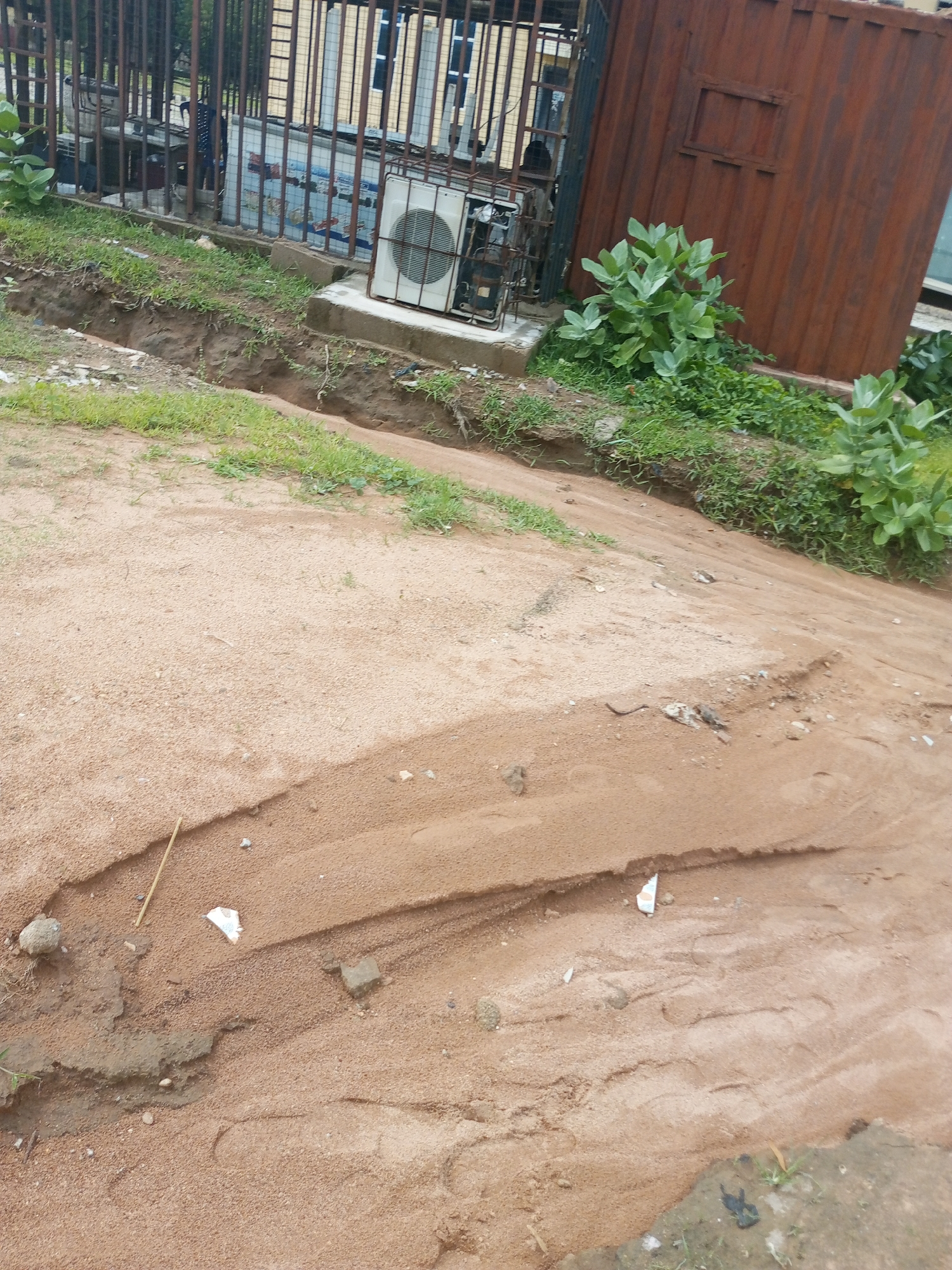 This media file captures an outdoor scene in Jalingo, Nigeria, likely during the day. The foreground is dominated by a sandy slope with visible erosion patterns, suggesting recent rainfall or water flow. Scattered across the sand are small rocks, debris, and a piece of white paper. A thin, dry stick lies diagonally across the sand.
In the midground, there's a trench or gully carved into the earth, with patches of green grass growing along its edges. Beyond the trench, a metal fence with vertical bars encloses an area. Inside this enclosure, several objects are visible, including what appears to be an air conditioning unit mounted on a concrete base, and a large, rusted corrugated metal container. Green leafy plants are growing near the fence and the container.
In the background, parts of a building with windows are visible behind the fence. There are no people clearly depicted in the scene, though figures may be present within the fenced area but are not distinct. The overall impression is one of an urban or semi-urban environment with signs of disuse or neglect. The weather appears overcast, contributing to the muted lighting. No legible text is visible in the image.